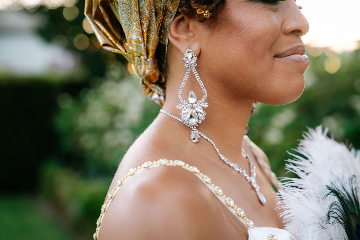 Close-up of a bride wearing elegant crystal drop earrings and a gold headwrap, with a white feather boa.