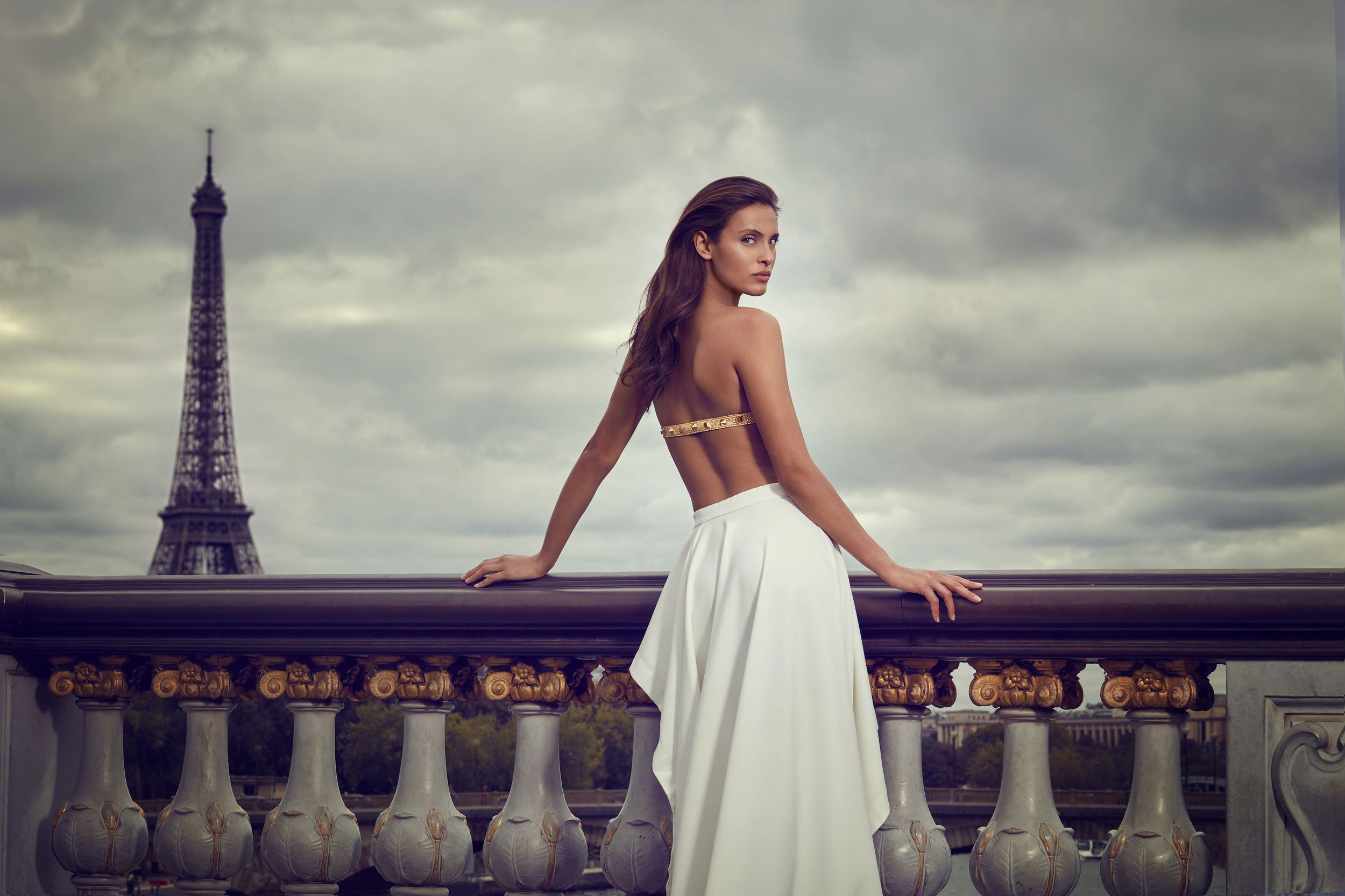 Elegant woman in white backless gown poses on Parisian balcony with Eiffel Tower in background.