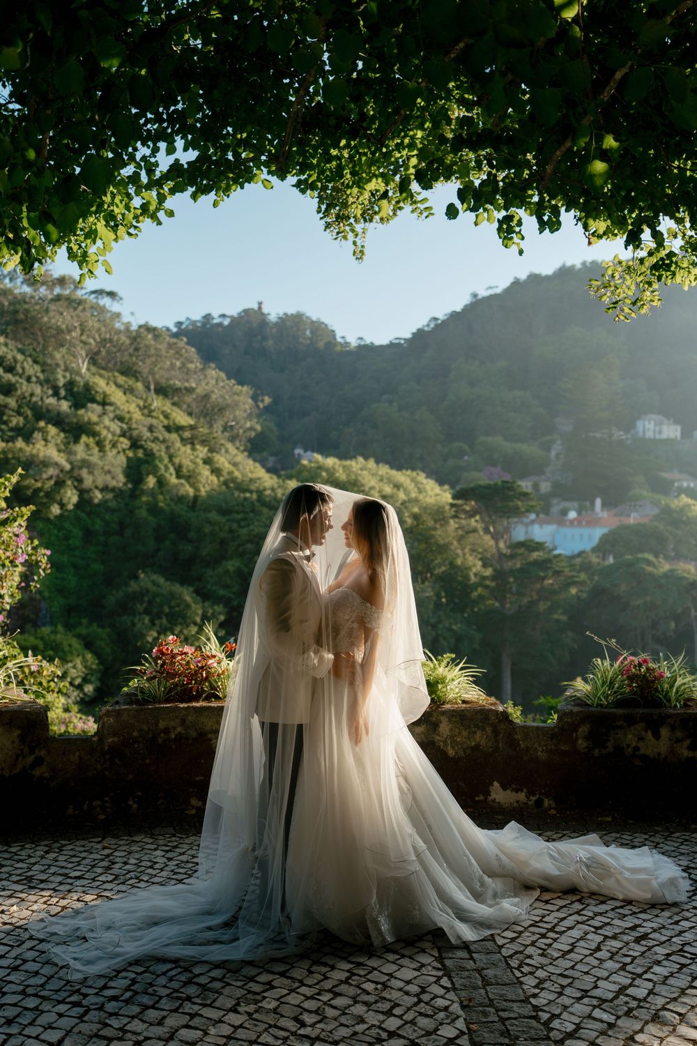 A couple in wedding attire share a romantic moment on a stone terrace overlooking lush mountain scenery at sunset.
