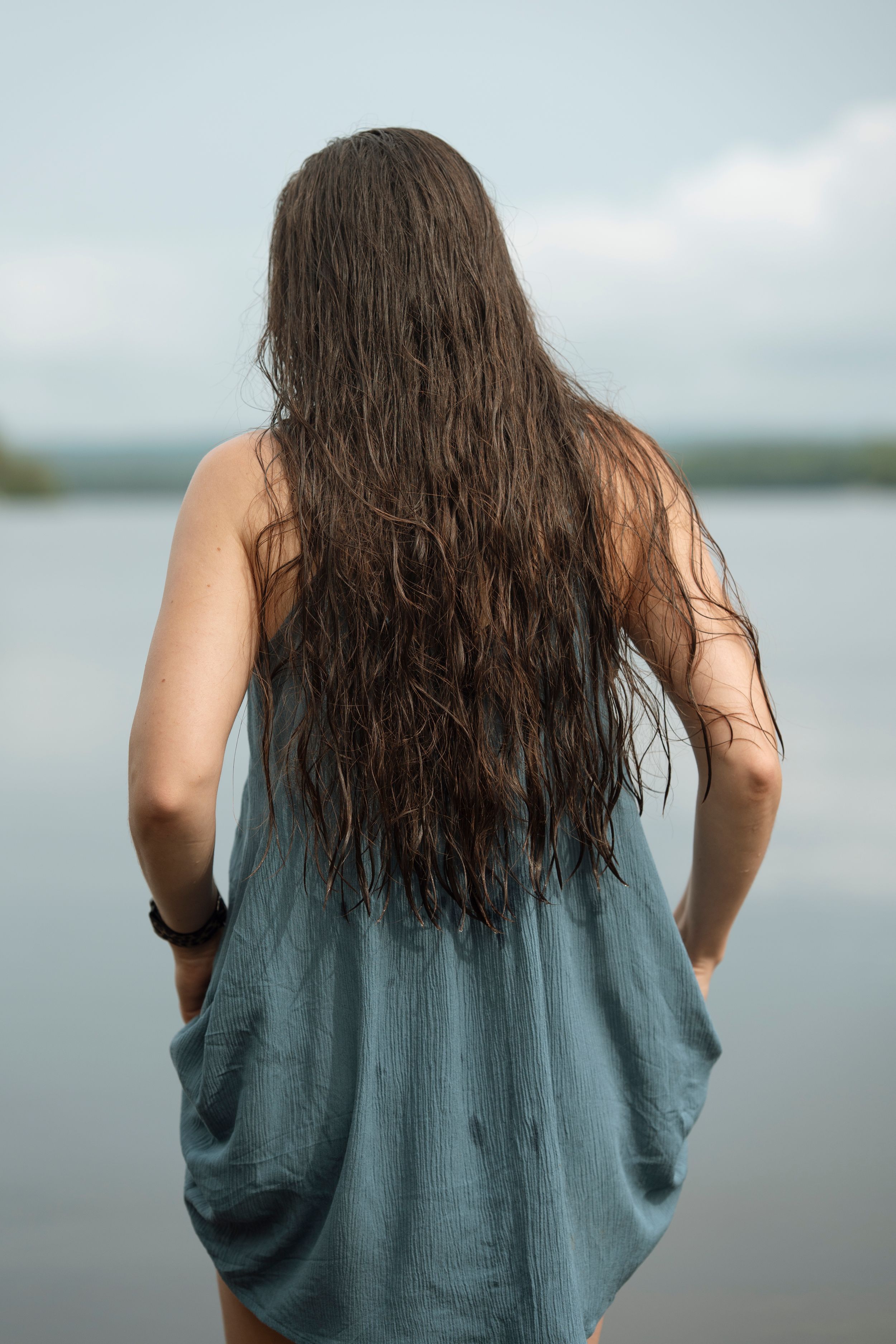 Woman with long wet dark hair wearing a teal dress stands facing a calm lake on a cloudy day.