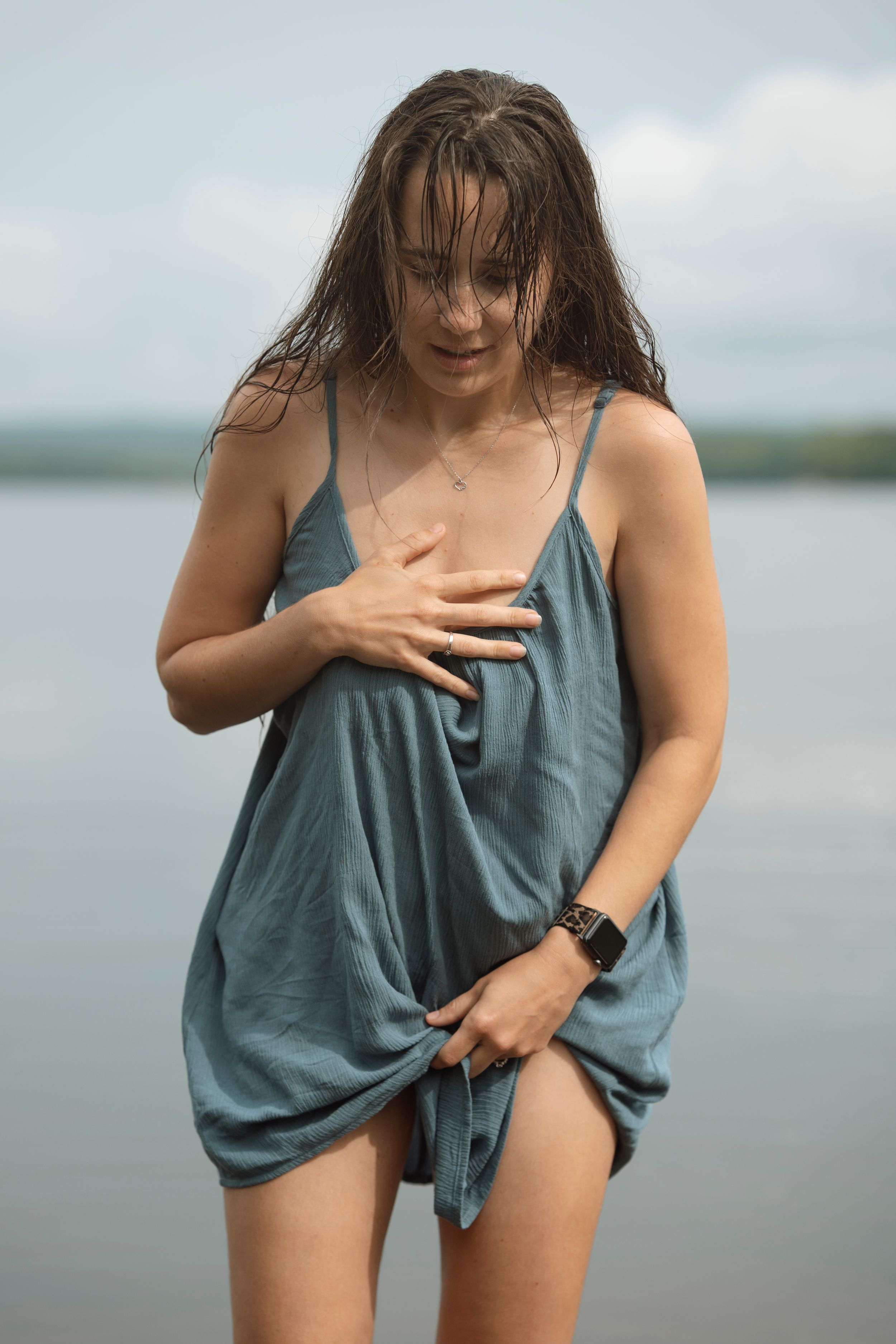 Young woman with wet hair wearing a teal slip dress standing by a lake on a cloudy day.