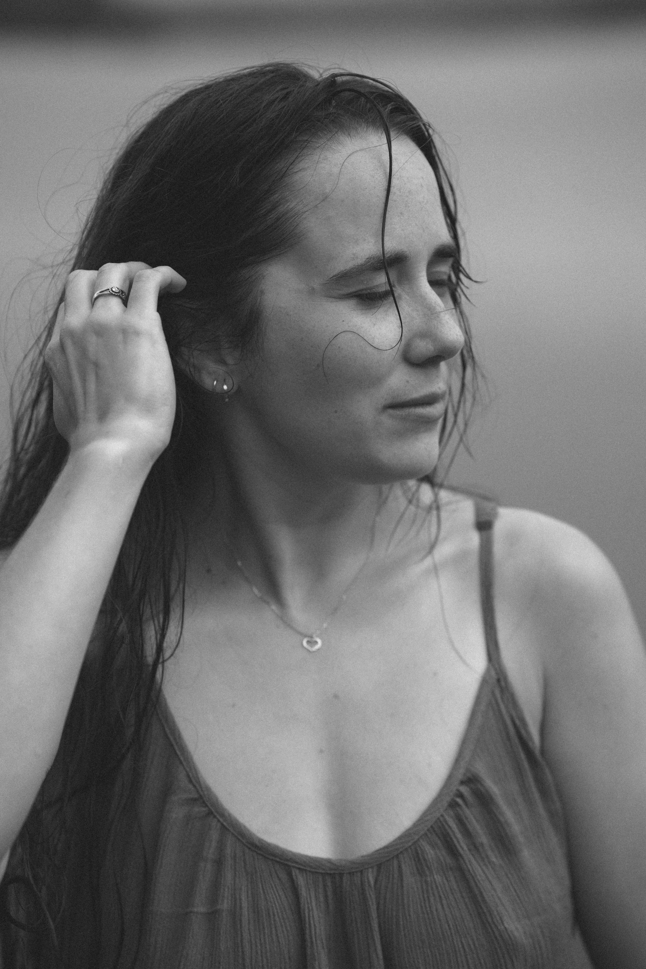 Black and white portrait of a young woman with wet hair, wearing a necklace and ring, eyes closed outdoors.