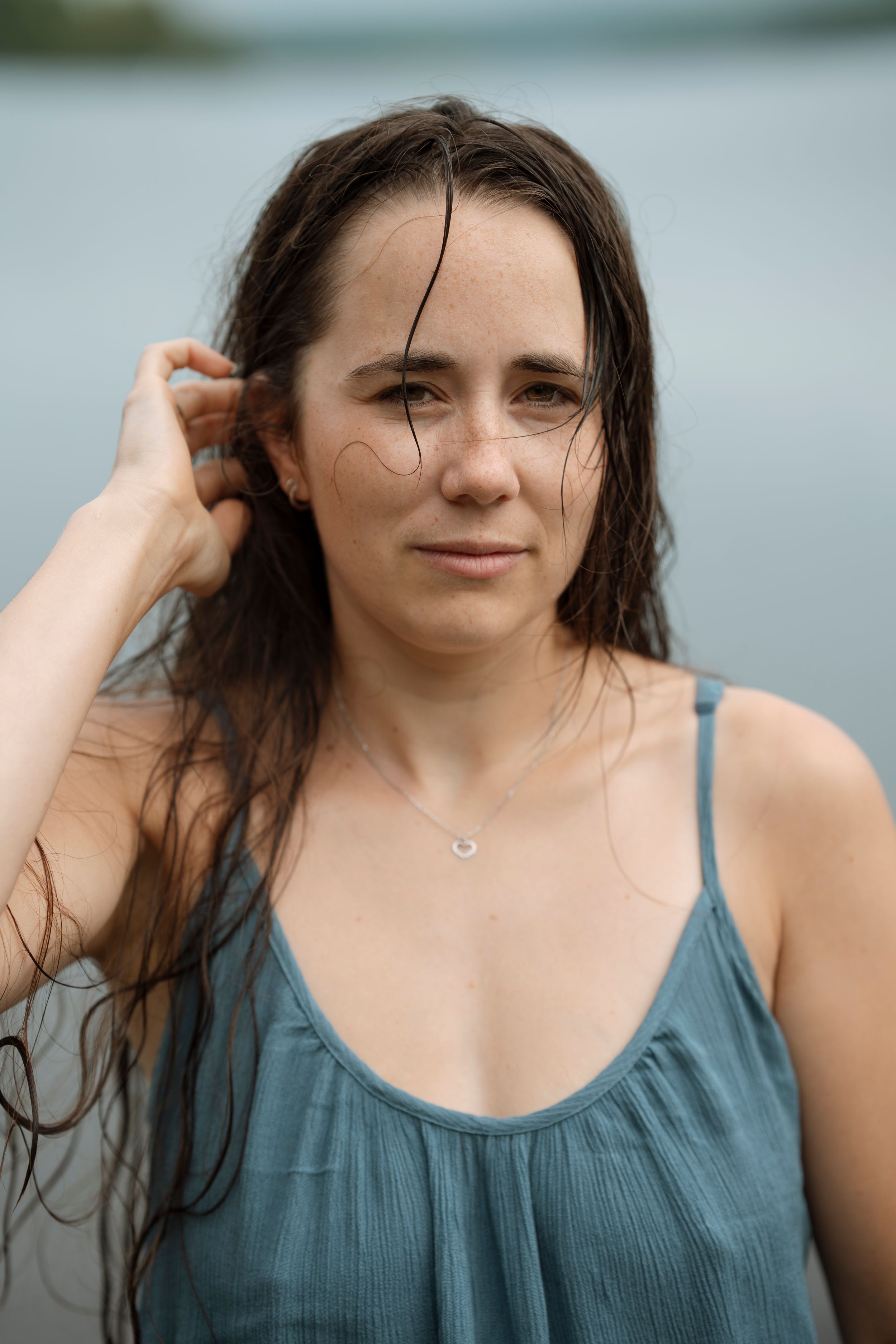 A young woman with wet dark hair poses near a lake, wearing a blue tank top and delicate necklace.
