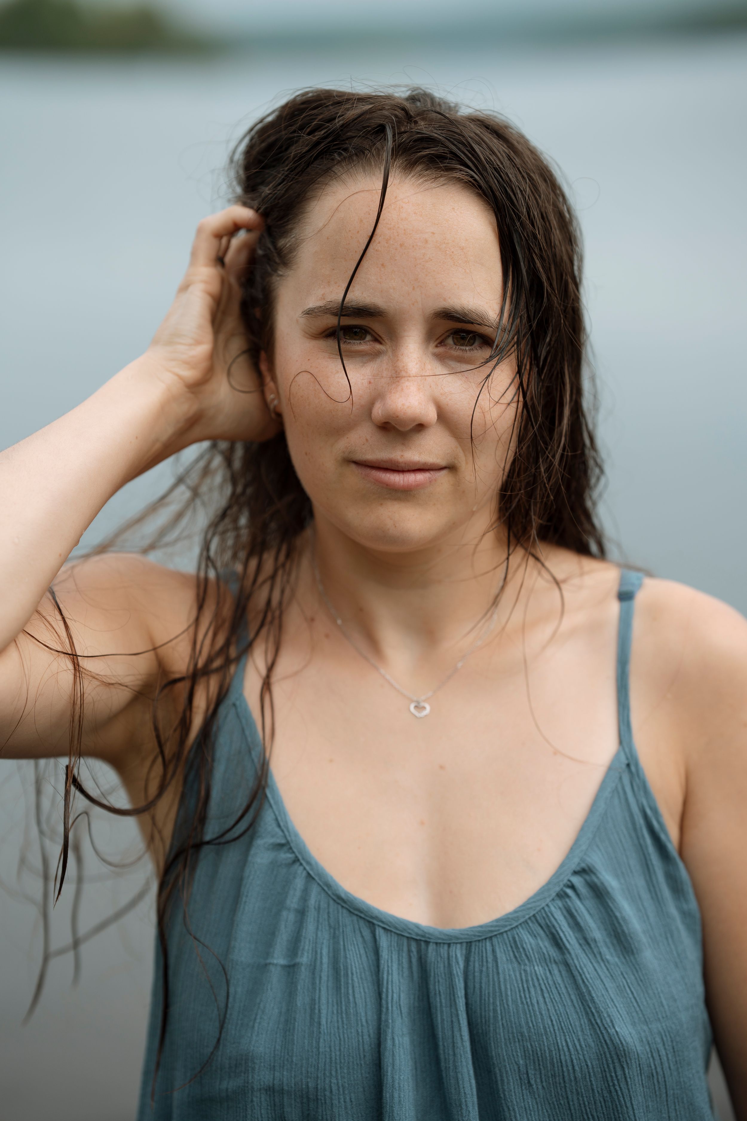 Woman with wet dark hair in teal sundress stands near water, hand in hair, wearing delicate necklace.