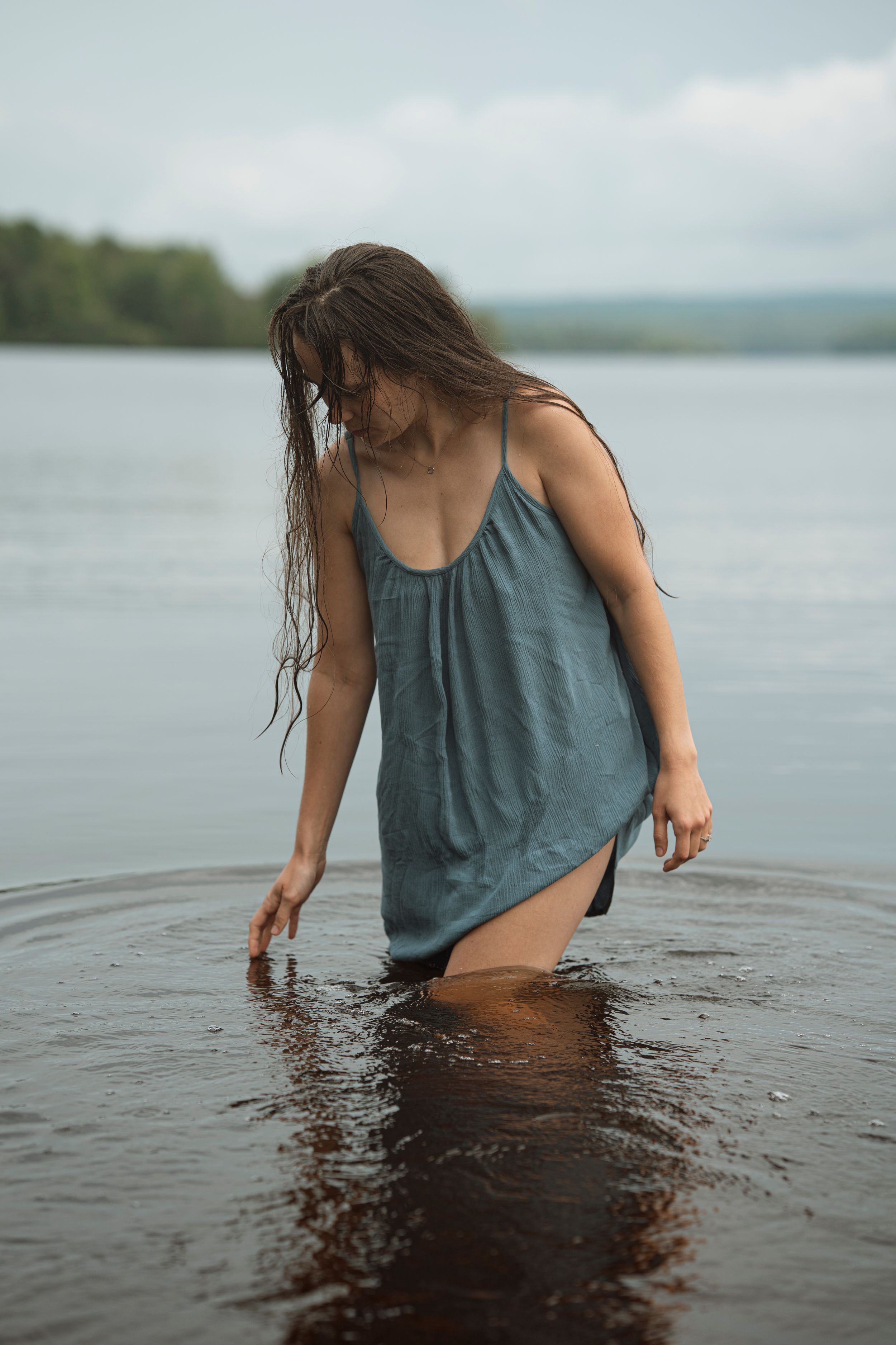 Woman in wet blue dress wading in calm lake water with long wet hair, misty forest background.
