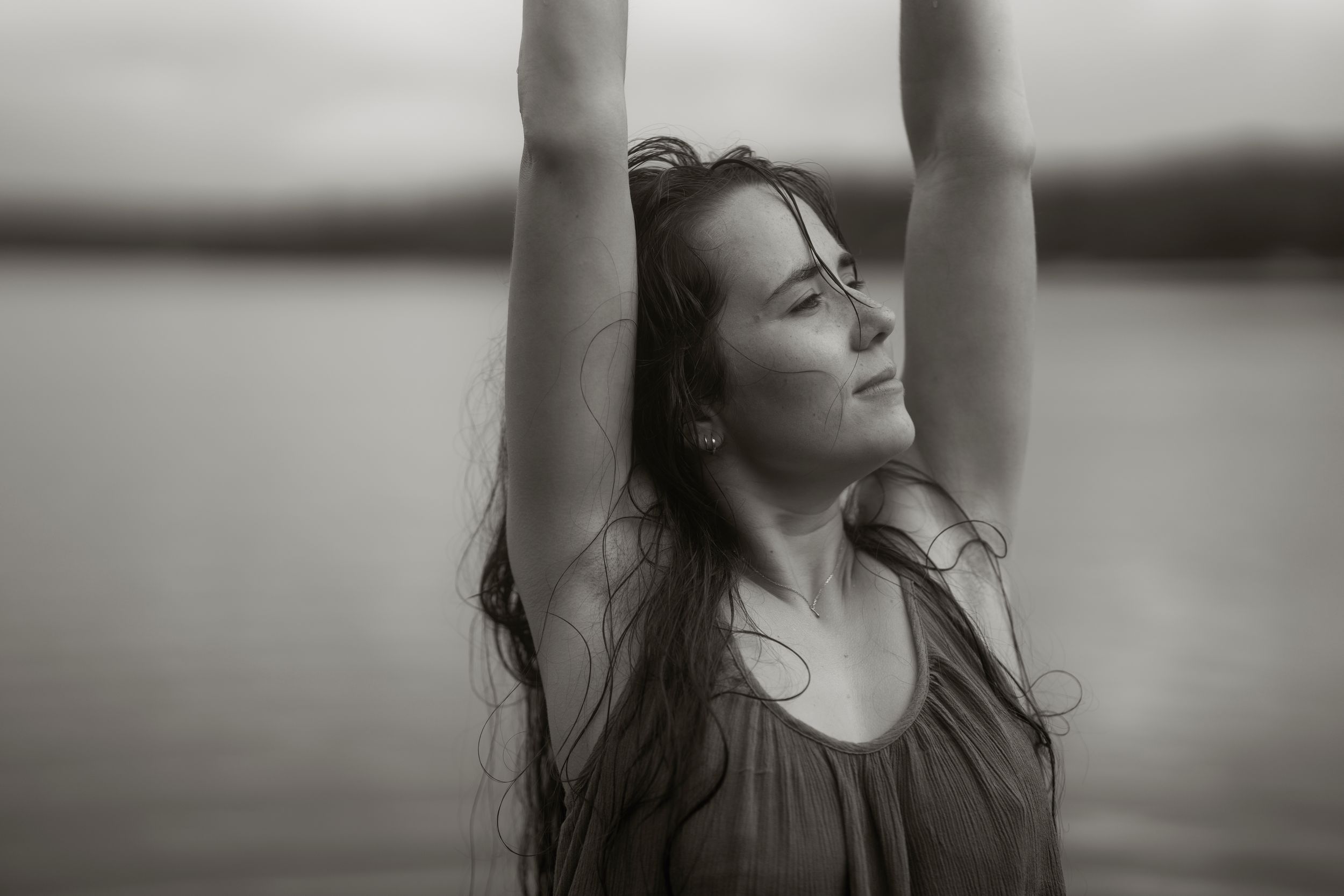 Black and white photo of a young woman with arms raised, eyes closed, long wet hair, standing near water.