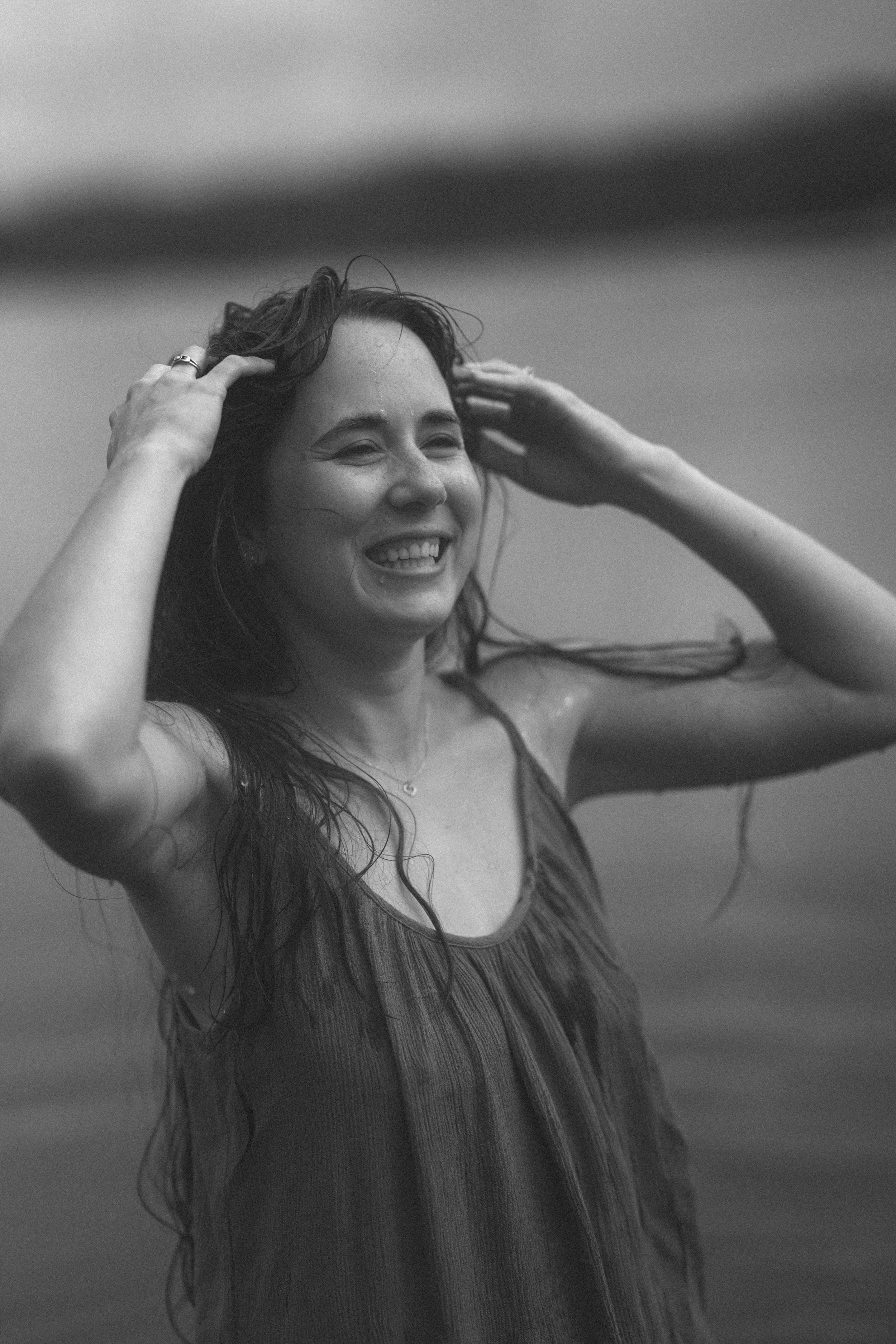 Black and white photo of a smiling woman with wet hair and dress, standing near water.
