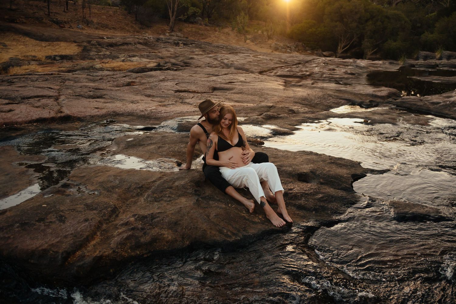 Expectant couple embraces beside flowing rock pools during a warm sunset maternity session at Roley Pools.