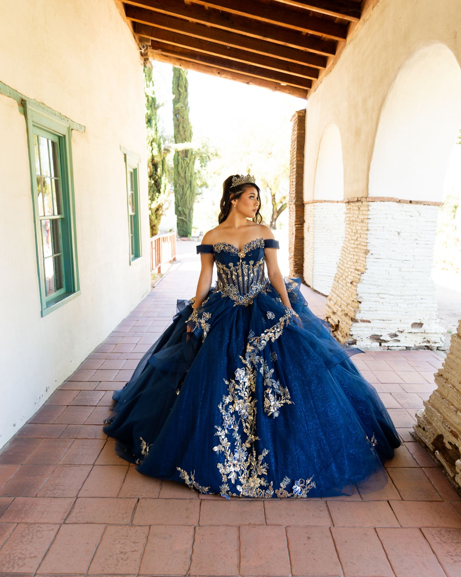 Young woman in navy blue quinceañera ball gown with gold floral embroidery poses in a rustic archway.