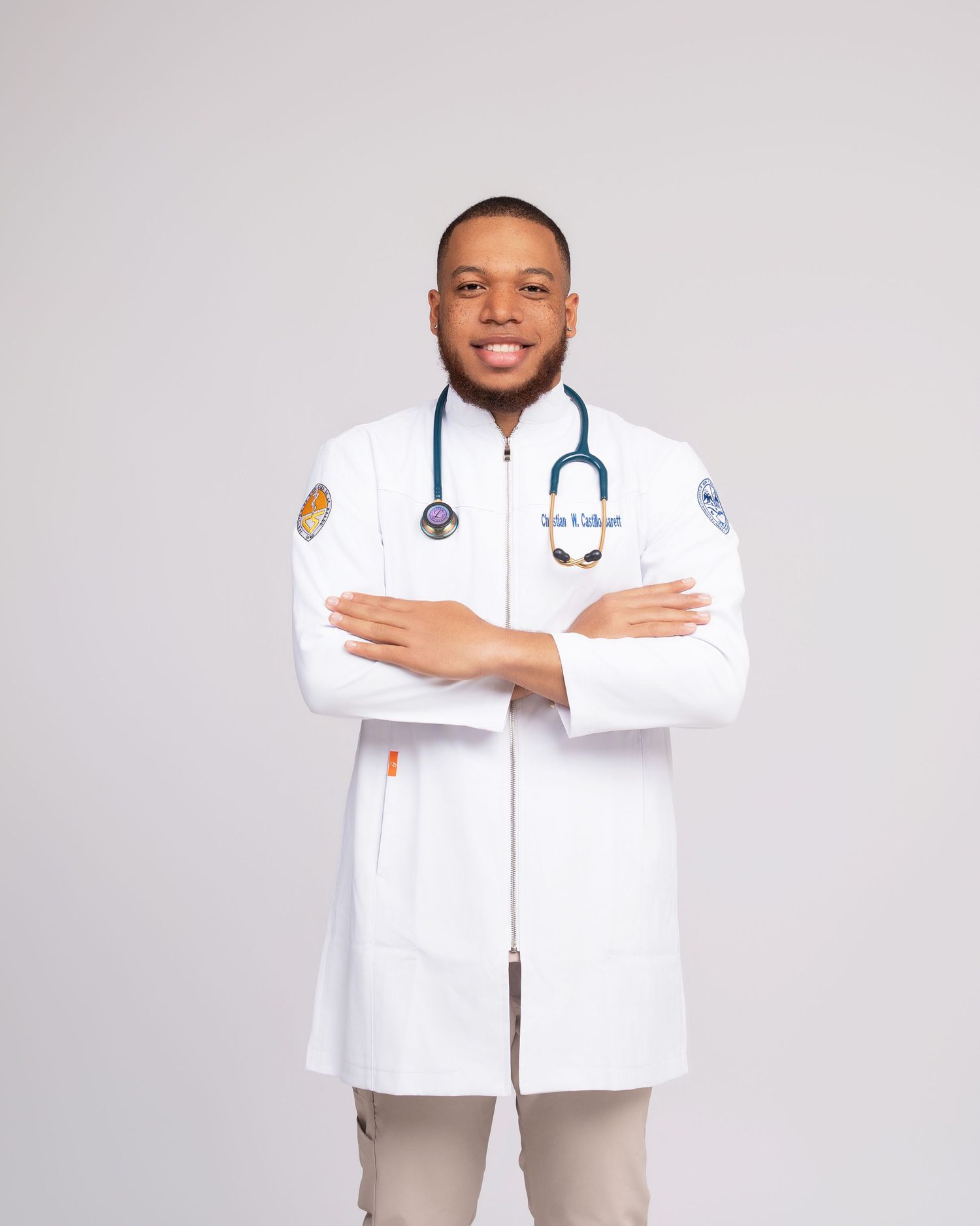 Woman in beige medical scrub jumpsuit poses with stethoscope beside black stool on gray background.