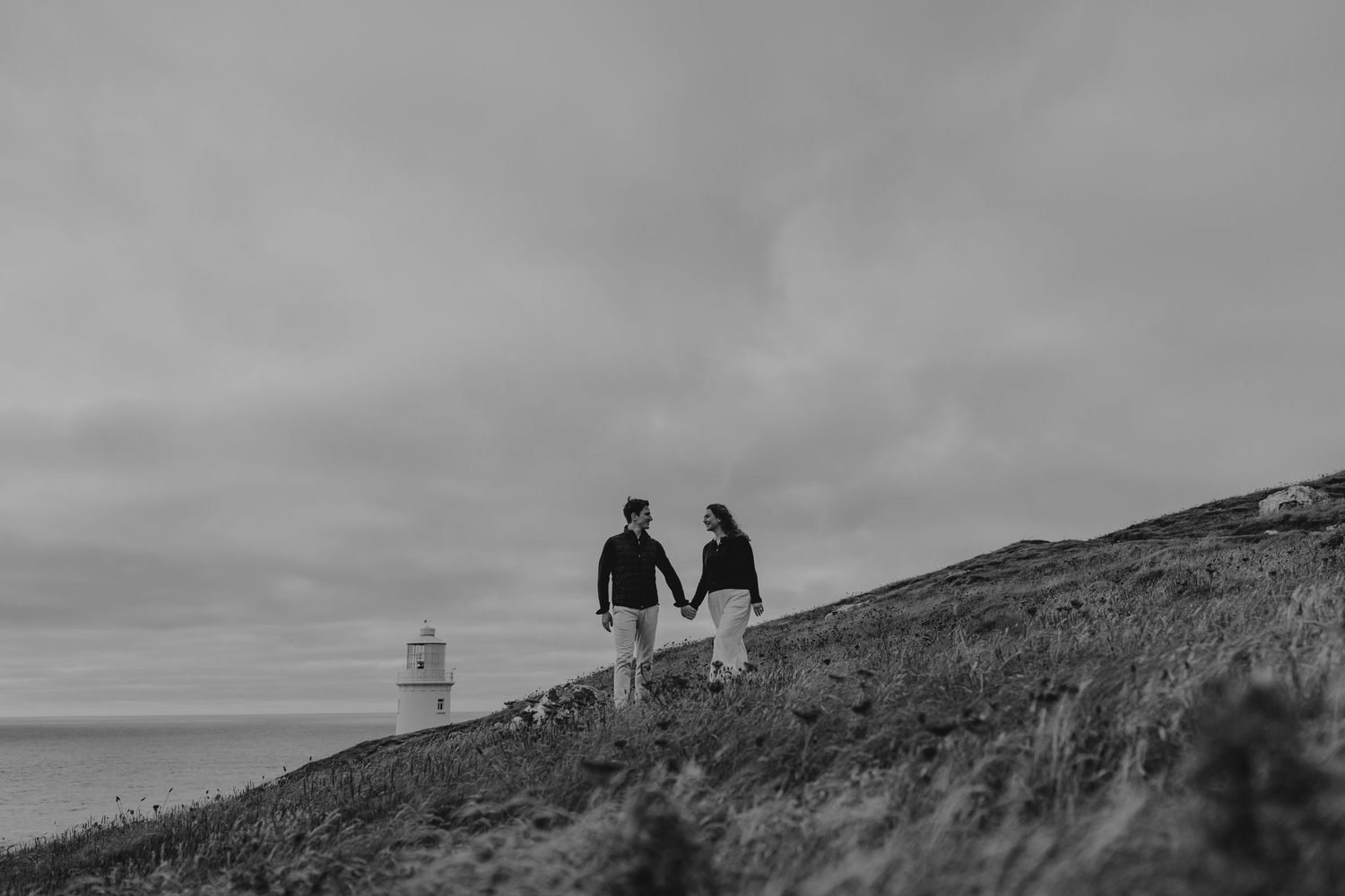 Black and white photo of a couple holding hands on a coastal hillside with a lighthouse in the background.