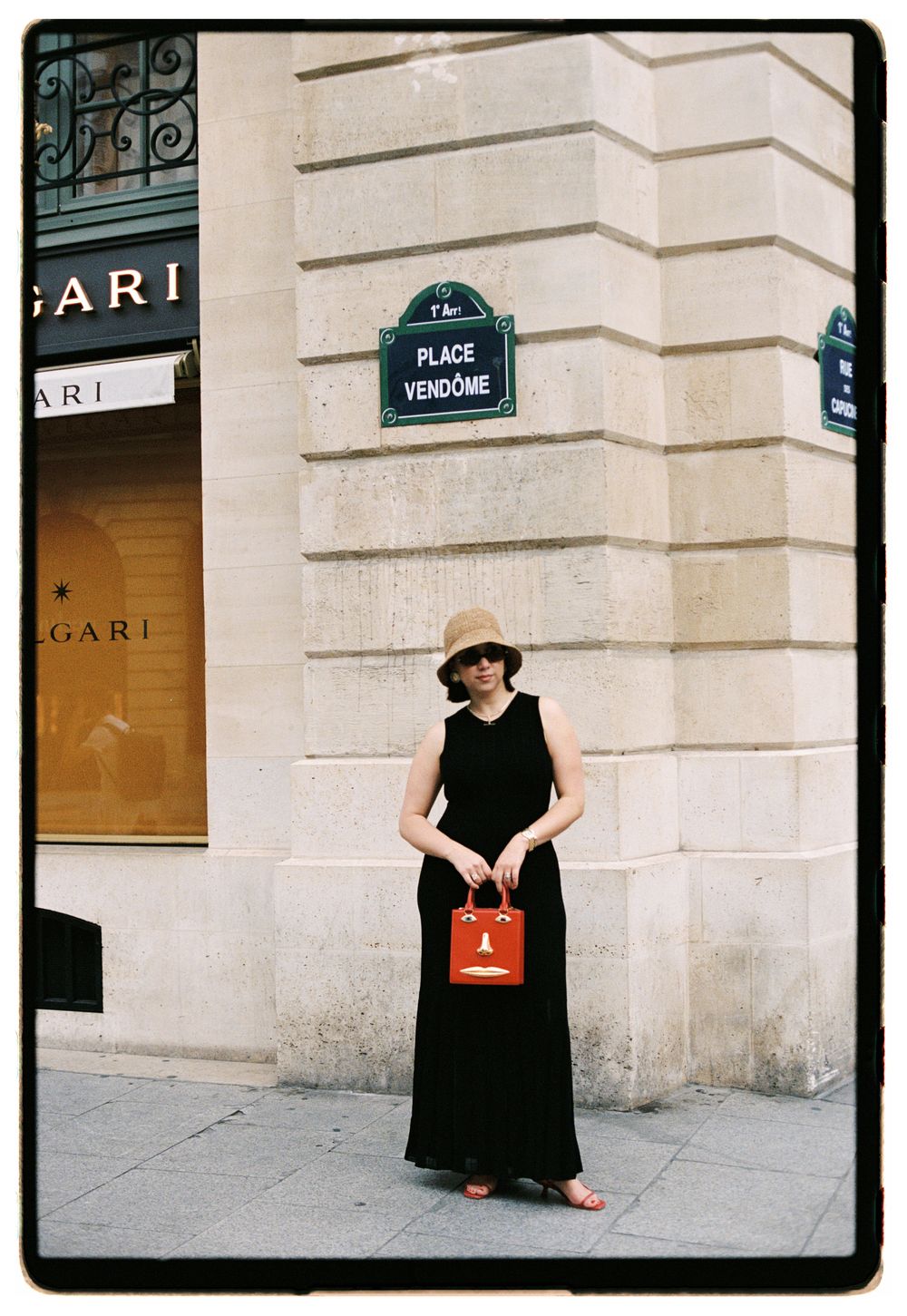 Woman in black maxi dress and sun hat holding red bag near Place Vendome sign in Paris. Schiaparelli photoshoot face bag, paris fashion