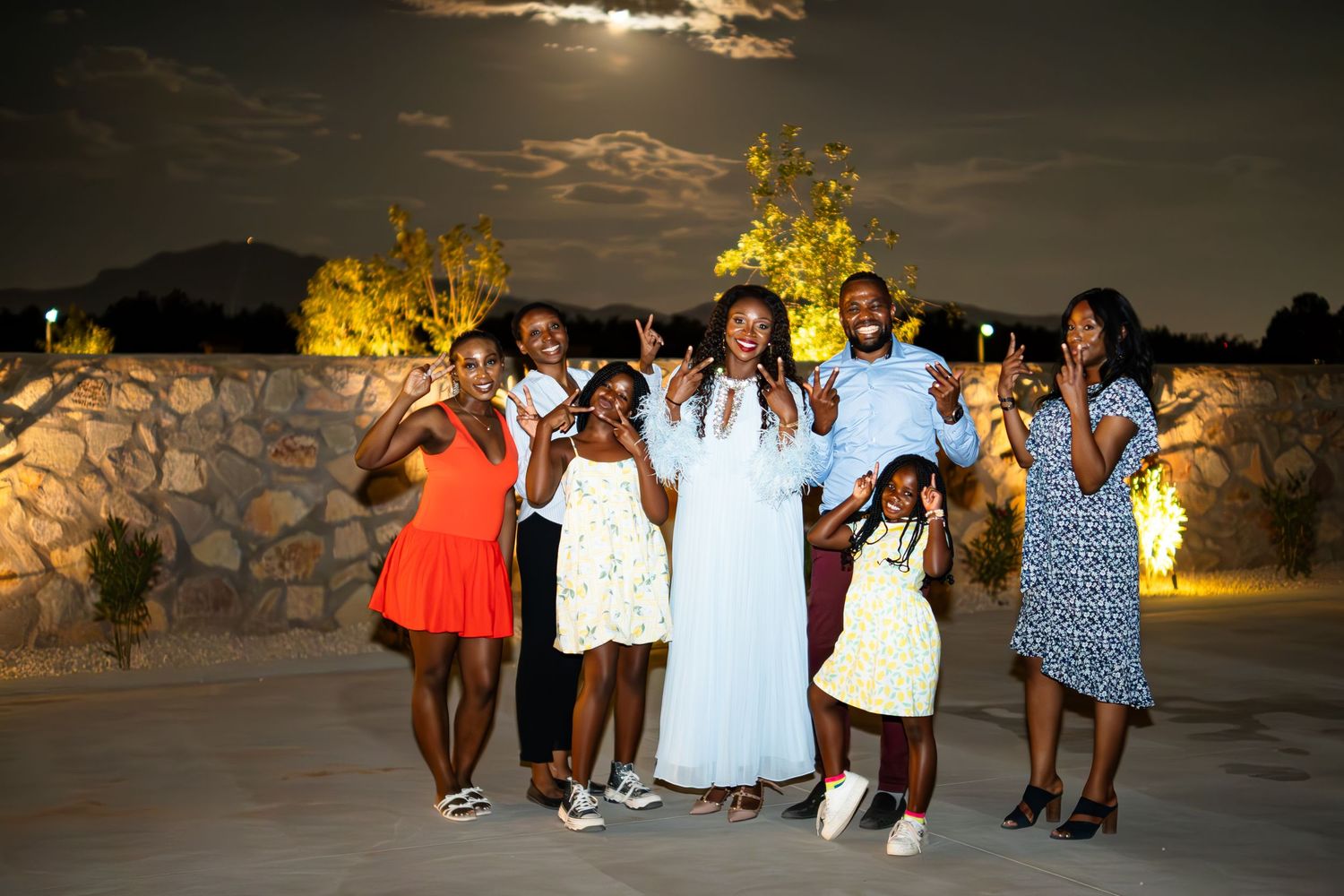 A joyful family of seven poses outdoors at night, making peace signs, with stone walls and warm lights behind them.