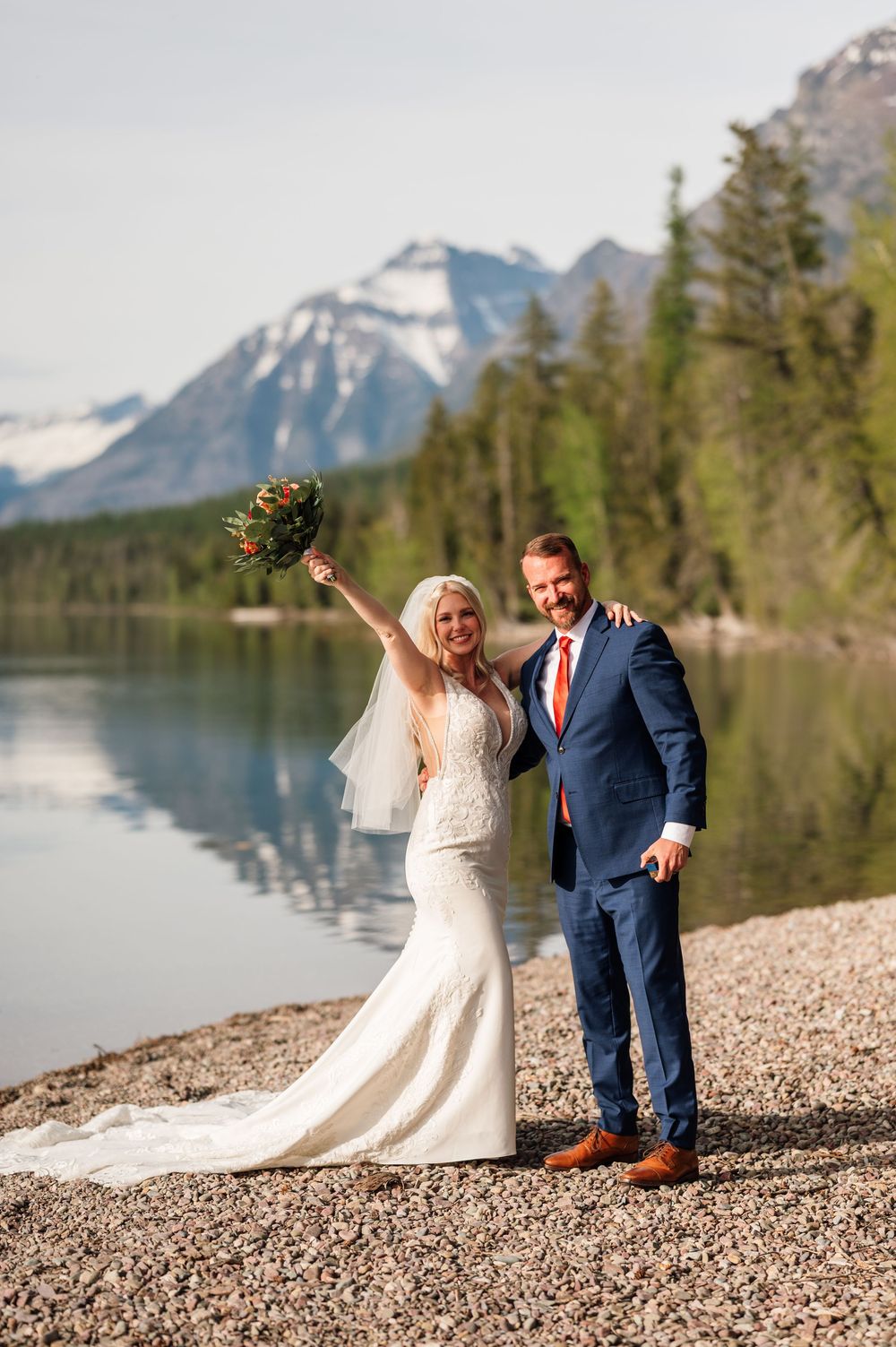 Bride and groom celebrate on a rocky lakeside with snow-capped mountains and pine trees in the background.