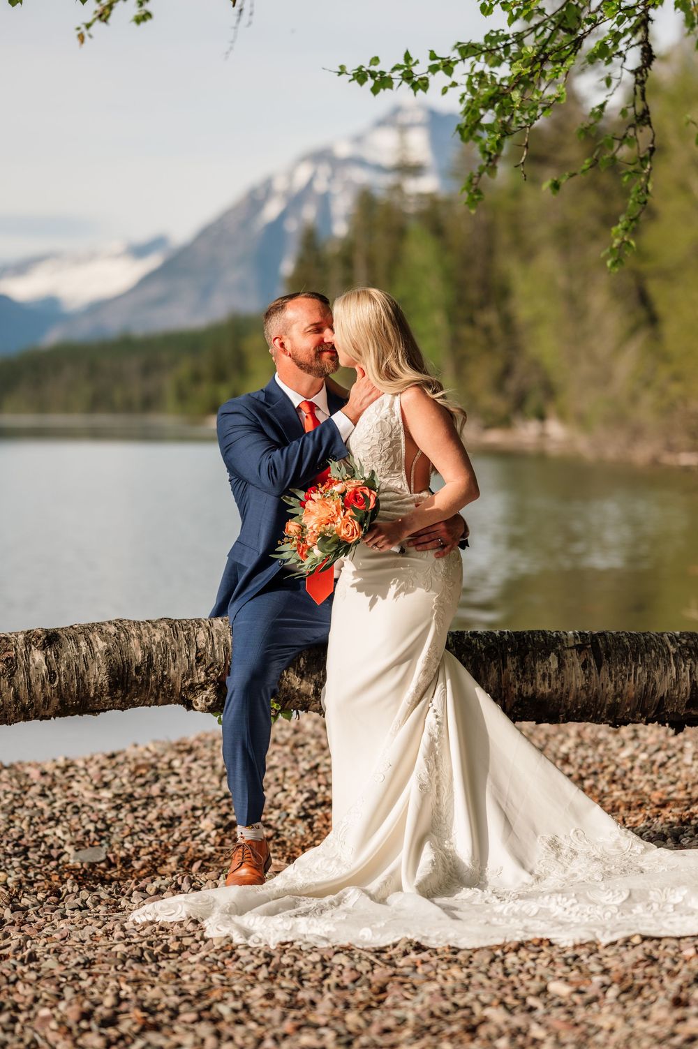Bride and groom share a romantic moment by a mountain lake, she holds a colorful bouquet, he wears a navy suit.