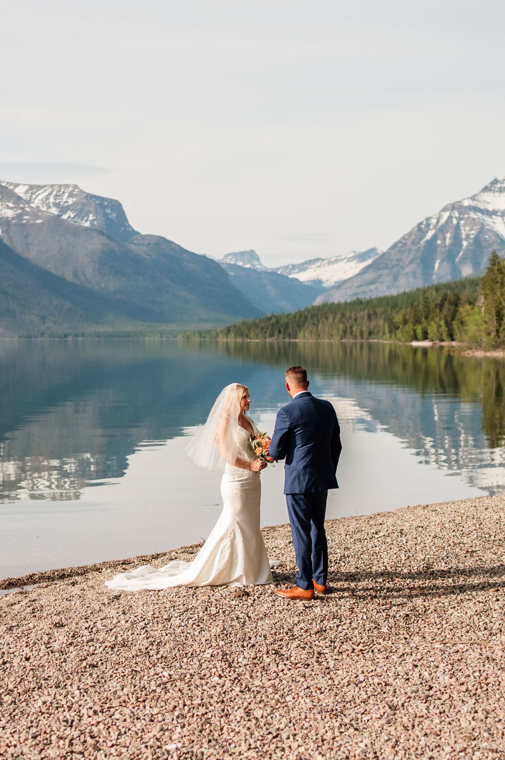 Bride and groom exchange vows on a rocky shoreline with a stunning mountain lake and snow-capped peaks backdrop.