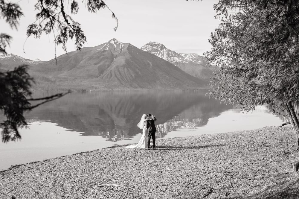 Black and white wedding photo of couple embracing on rocky lakeshore with mountain reflection in calm water.