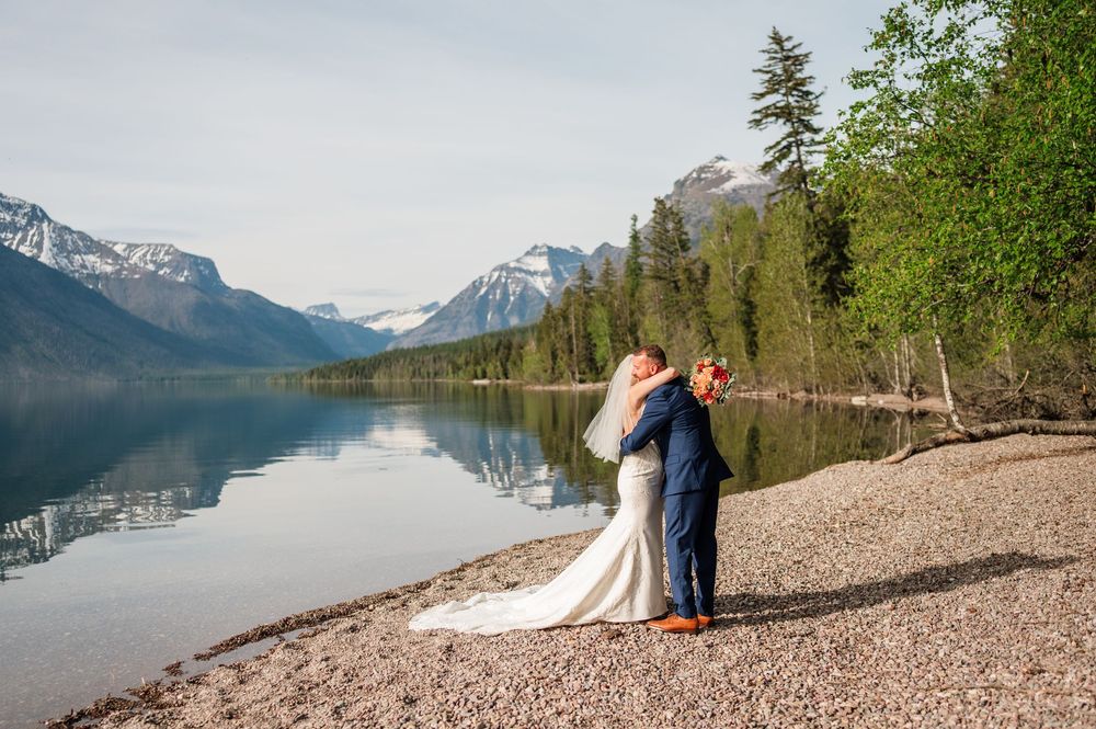 Bride and groom embrace on a rocky lakeshore with stunning mountain reflections and evergreen trees at Glacier National Park.