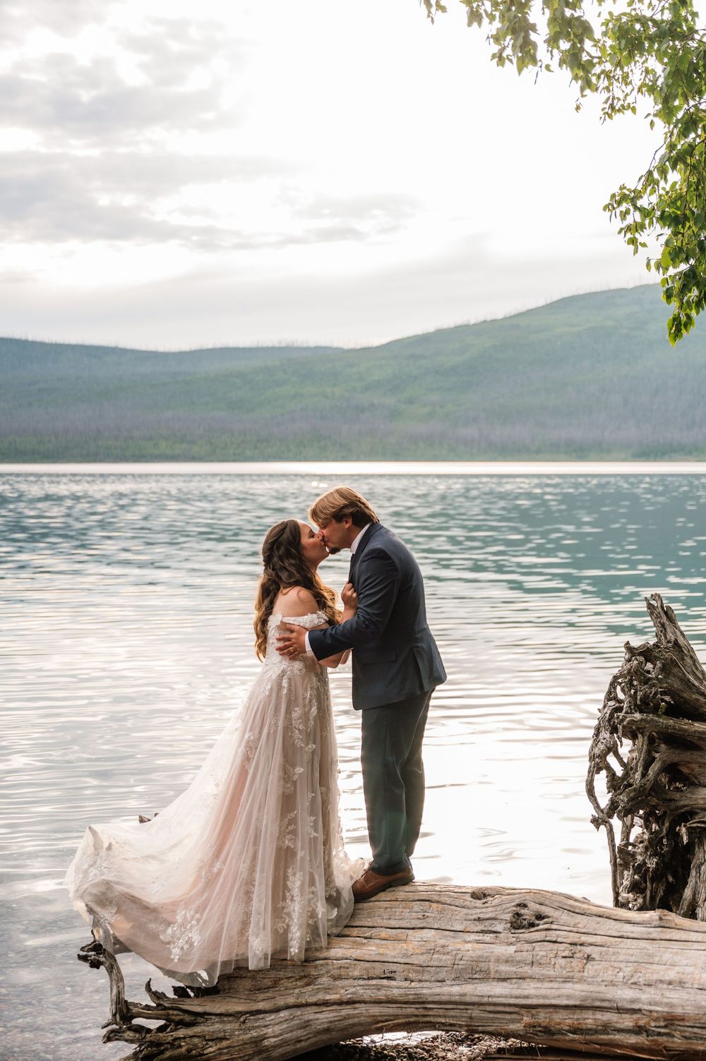 Bride and groom share a romantic kiss on a log by a serene mountain lake in a stunning outdoor wedding setting.