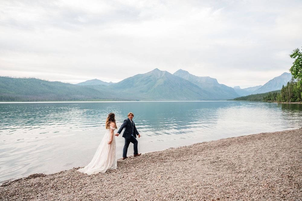 Couple in wedding attire walks hand-in-hand along a rocky lakeshore with misty mountains in the background.