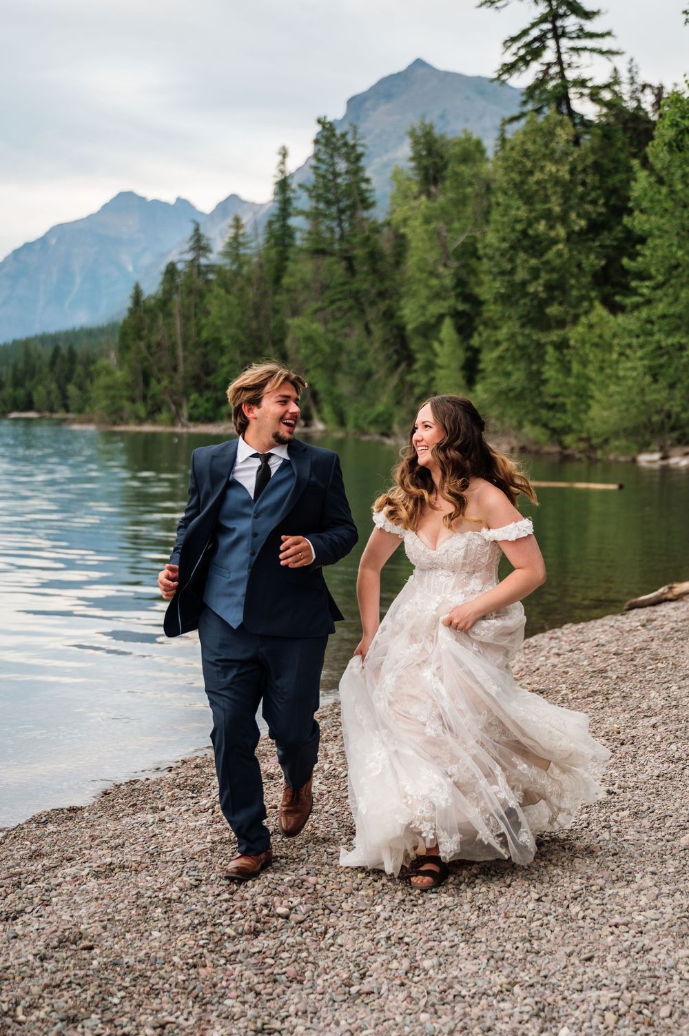 A joyful bride and groom run along a rocky lakeside shore with mountain forest backdrop on their wedding day.