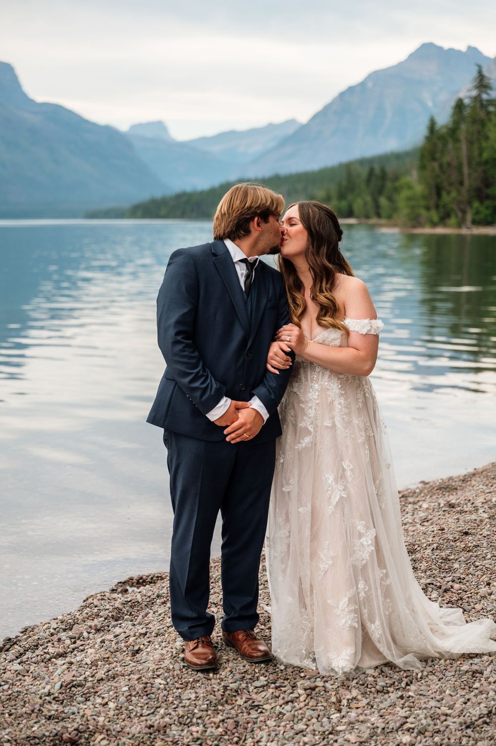 A bride and groom share a kiss by a serene mountain lake, she in a floral gown, he in a navy suit.