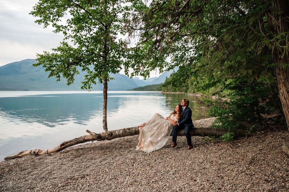 Bride and groom embrace near a fallen tree on a rocky lakeshore with mountains and calm water in the background.