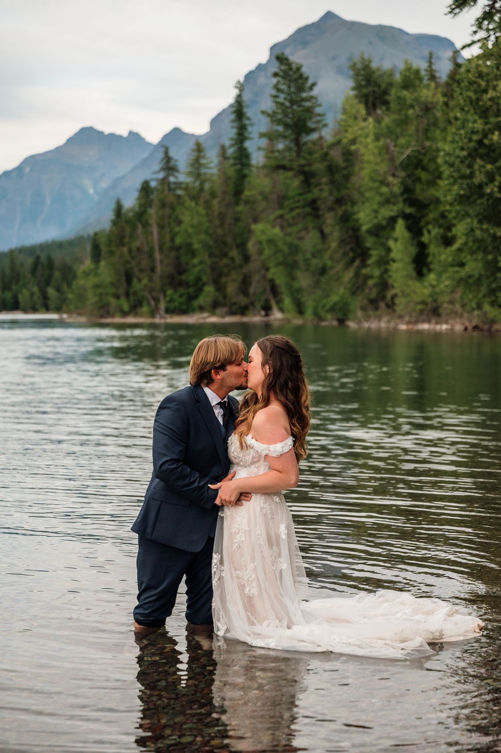 Bride and groom kissing in a mountain lake wearing wedding attire, surrounded by pine trees and rocky peaks.