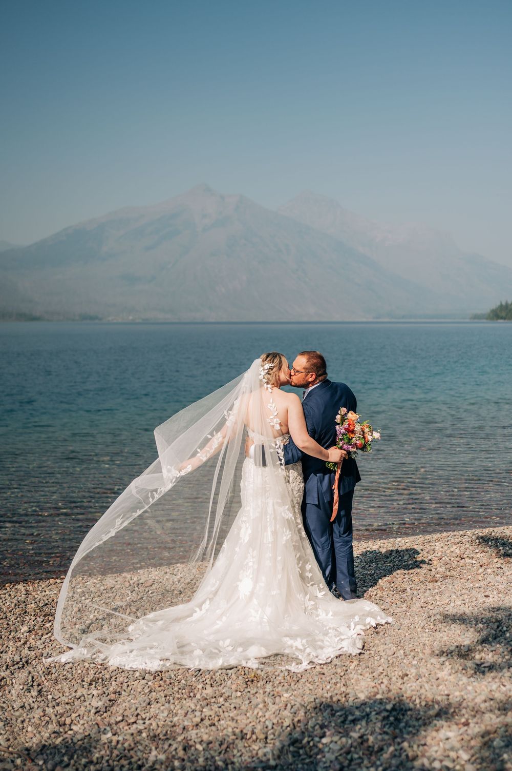 Bride and groom share a kiss by a mountain lake, her veil flowing in the wind, wearing white lace gown and navy suit.