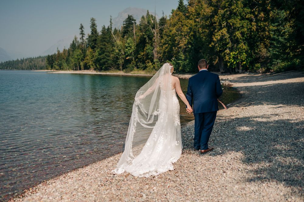 Bride and groom walk hand-in-hand along a rocky lakeside shore surrounded by lush green forest on their wedding day.