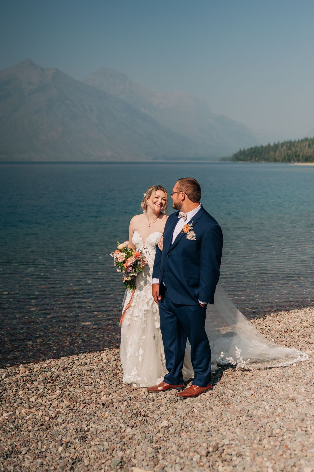 Bride and groom share a romantic moment on a rocky lakeside shore with mountain views on their wedding day.