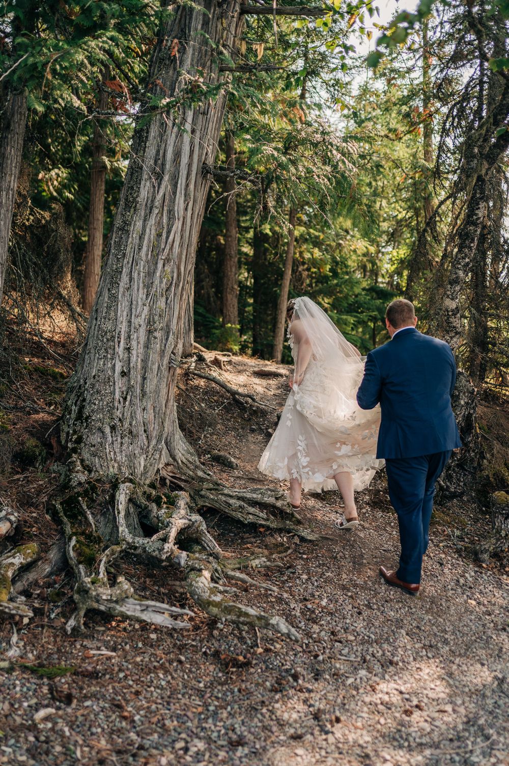 Bride and groom walking through a lush forest trail, her white gown and veil flowing as he holds it gently.