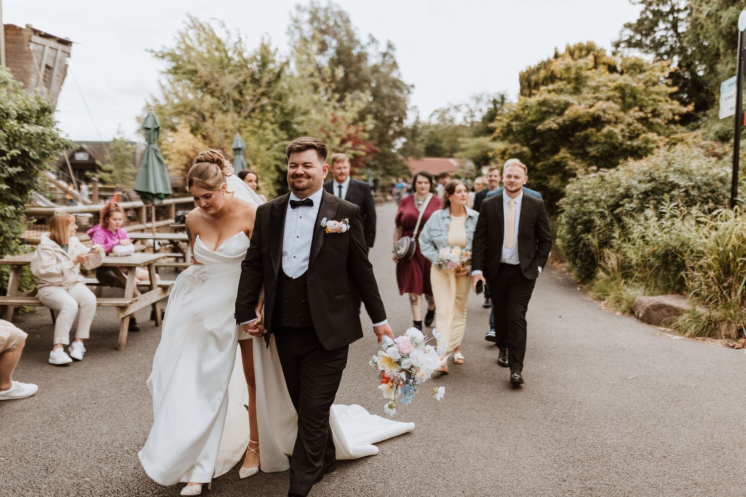 Happy newlyweds walk hand-in-hand at outdoor wedding venue surrounded by guests and lush greenery.