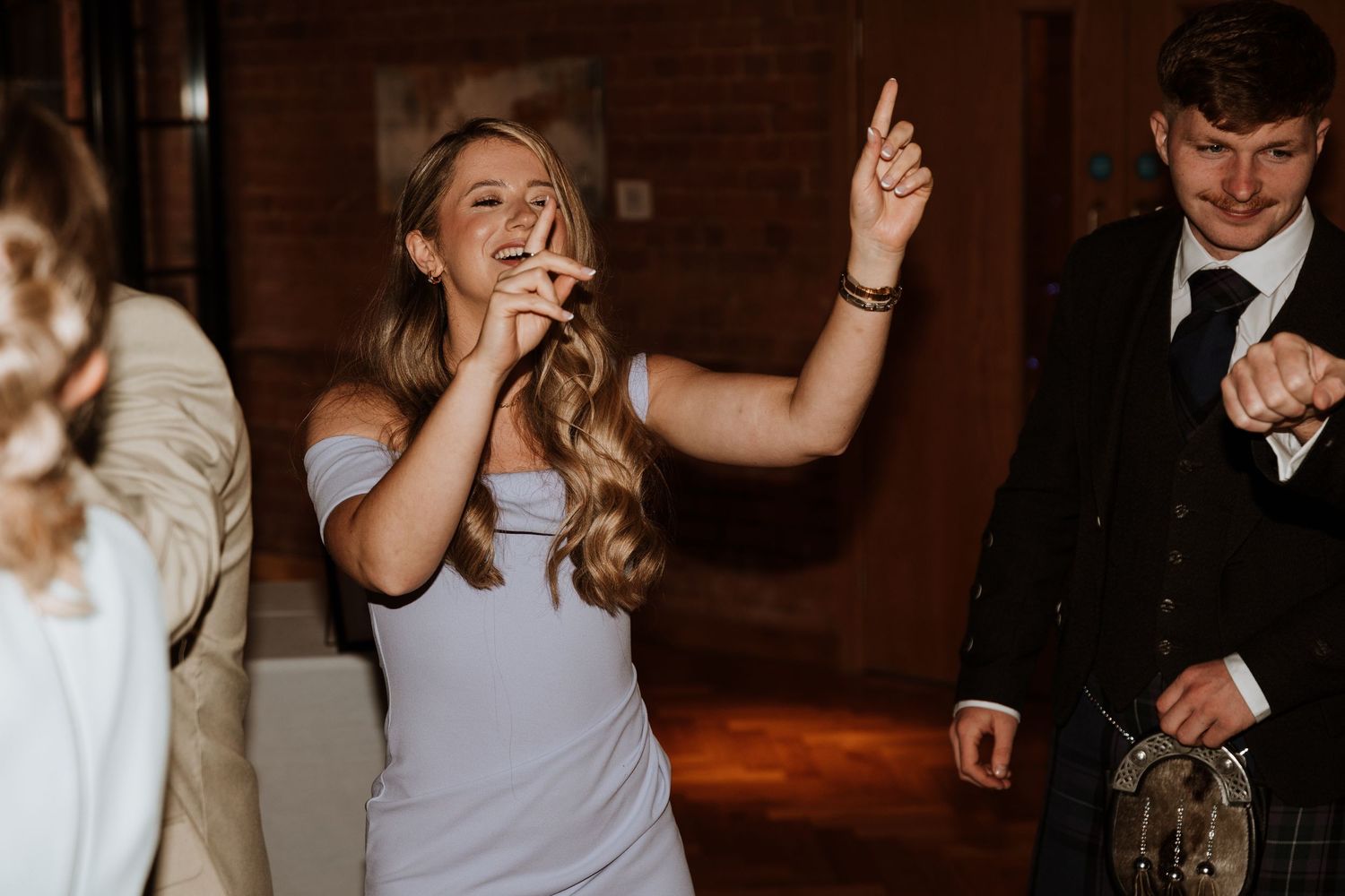 Woman in blue dress dancing and pointing up at a wedding reception with guests in kilts nearby.