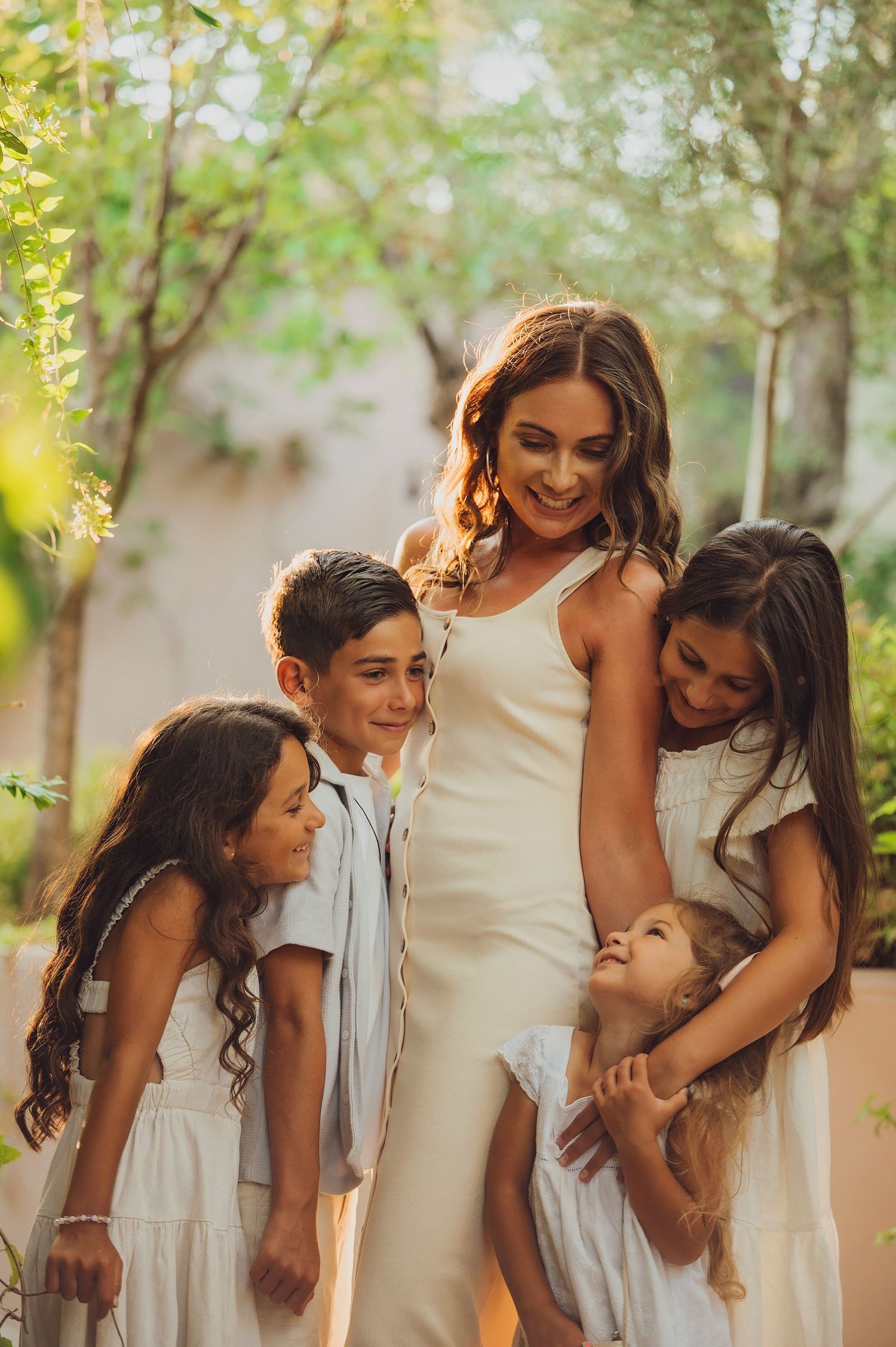A smiling woman in a white dress surrounded by four happy children in white outfits, posing outdoors among lush green trees.