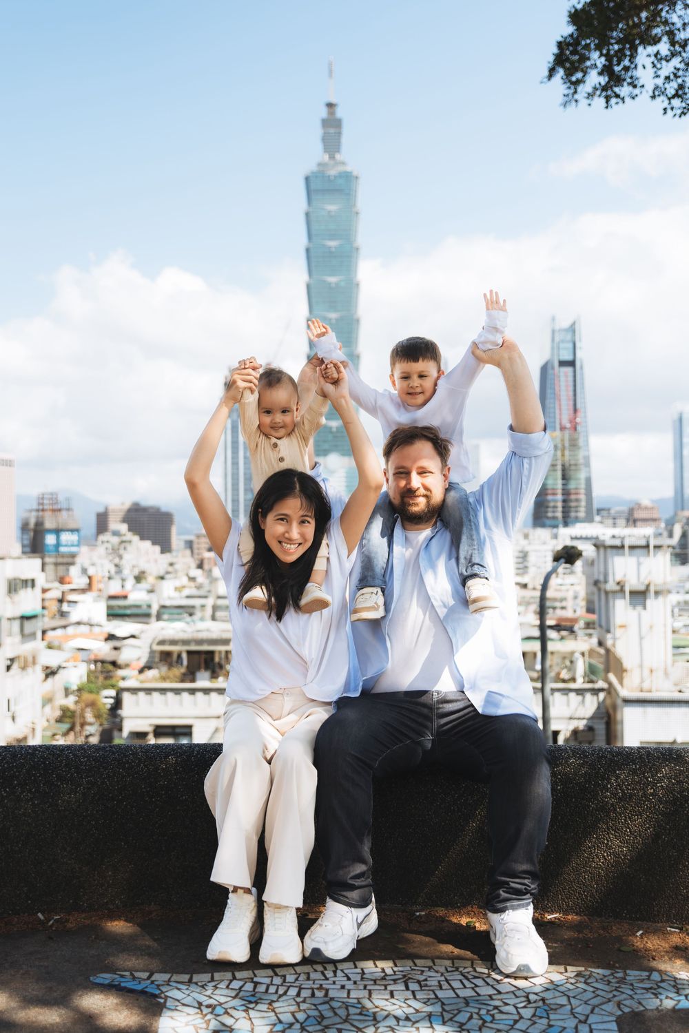 Happy family of four posing with Taipei 101 skyline in the background on a sunny day.