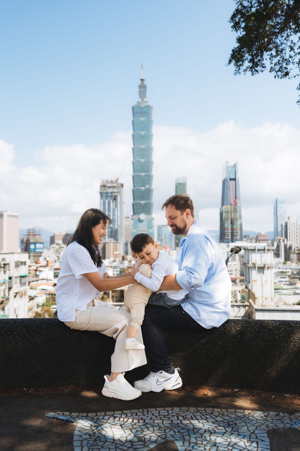 Family with baby posing on wall with Taipei 101 skyscraper skyline in background on sunny day.