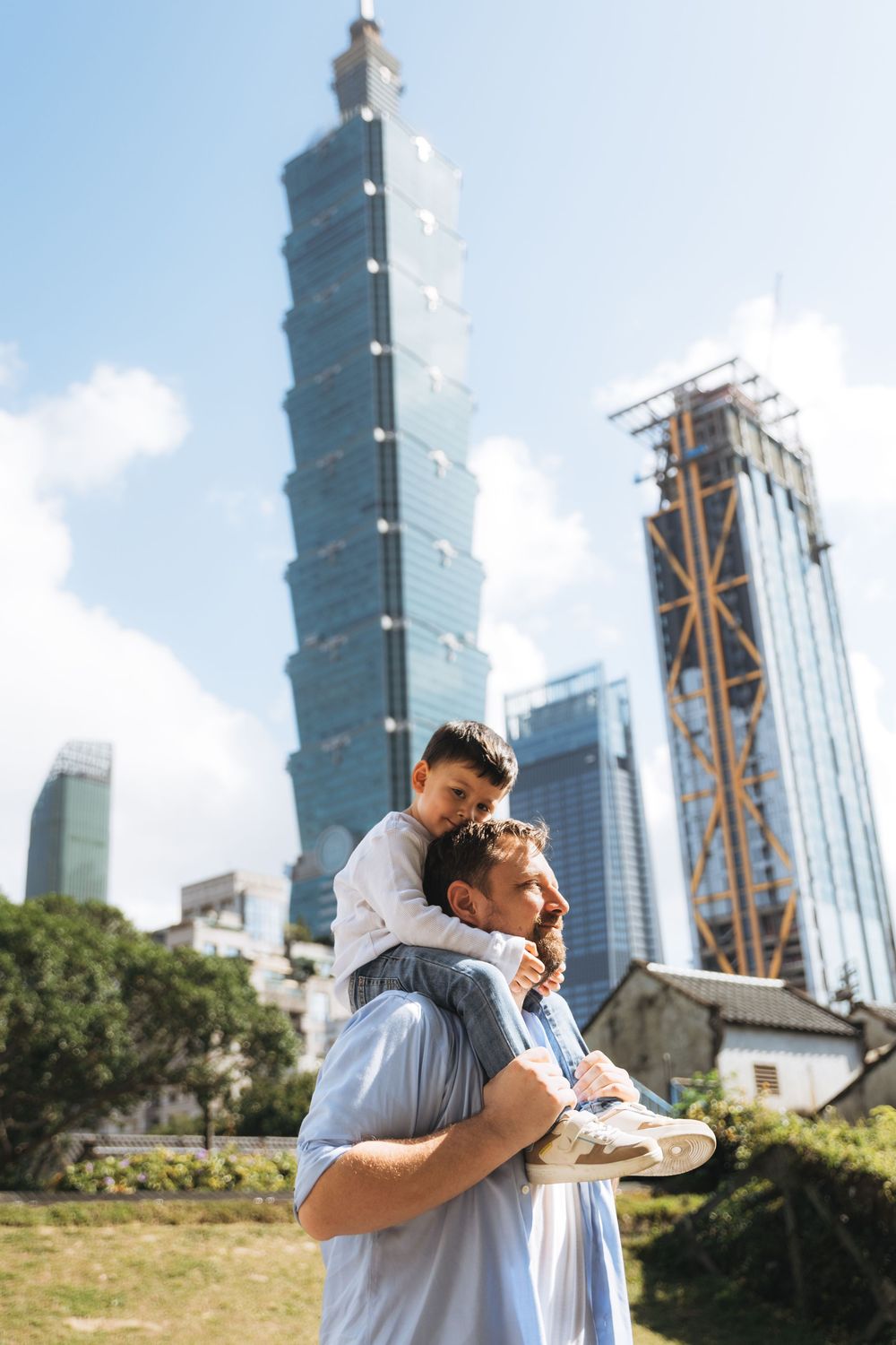 Father carries child on shoulders near Taipei 101 skyscraper on a sunny day in Taiwan.
