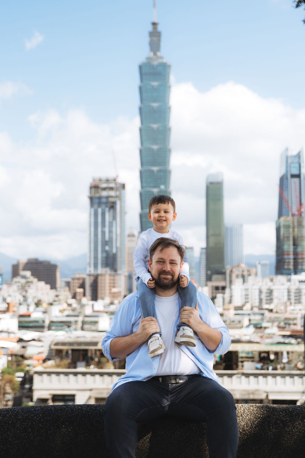 Father and son posing with Taipei 101 skyscraper skyline in background on a sunny day.