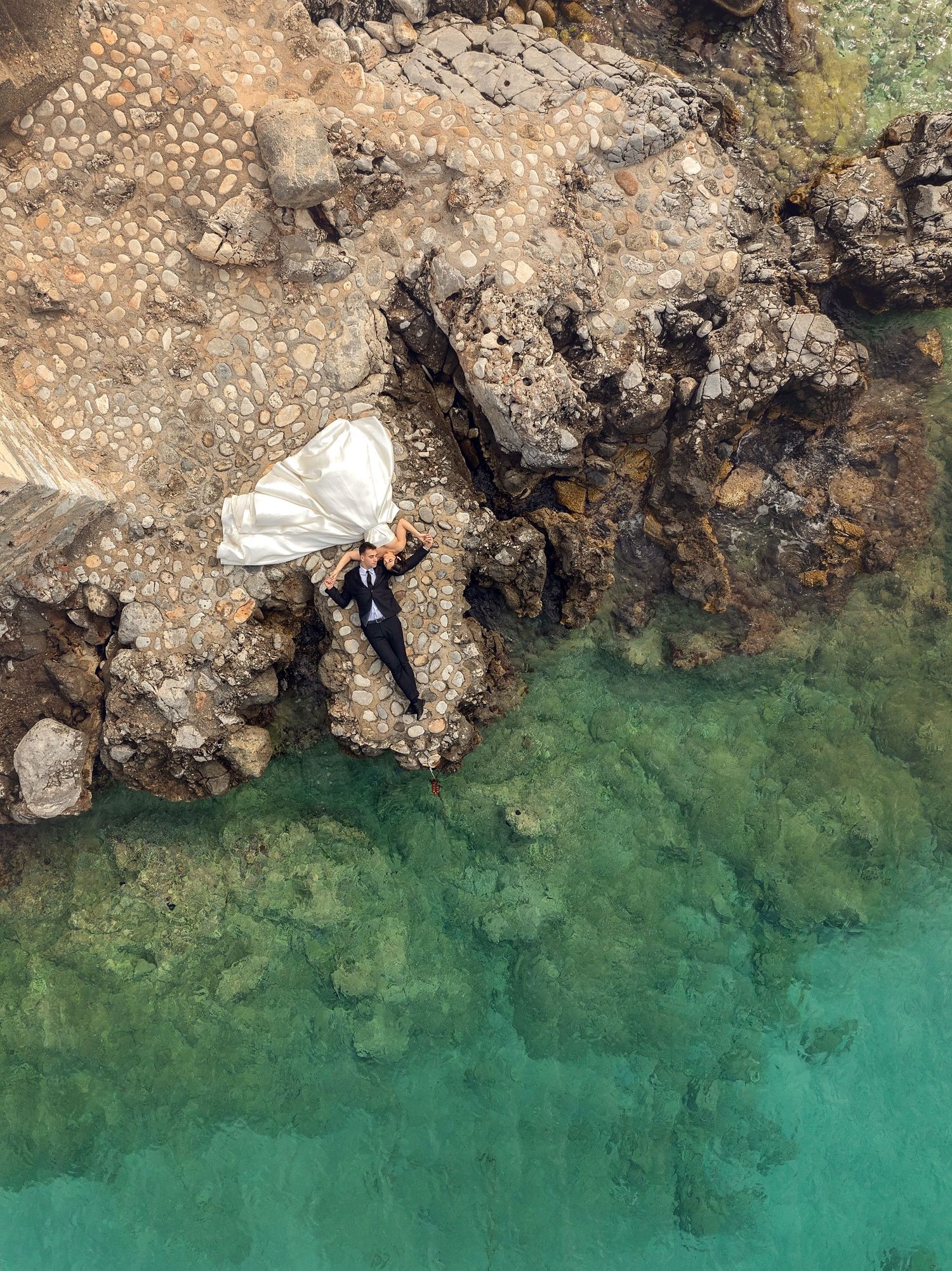 Aerial drone view of bride and groom lying on rocky coastal cliffs beside turquoise ocean waters.