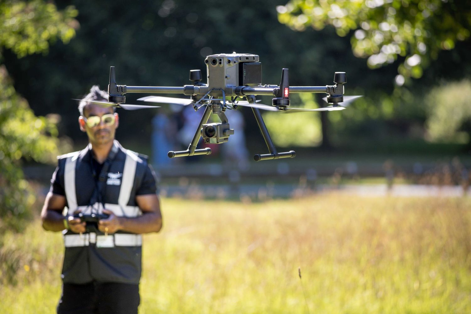 A drone operator pilots a large multi-rotor drone outdoors in a sunny green field using a remote controller.