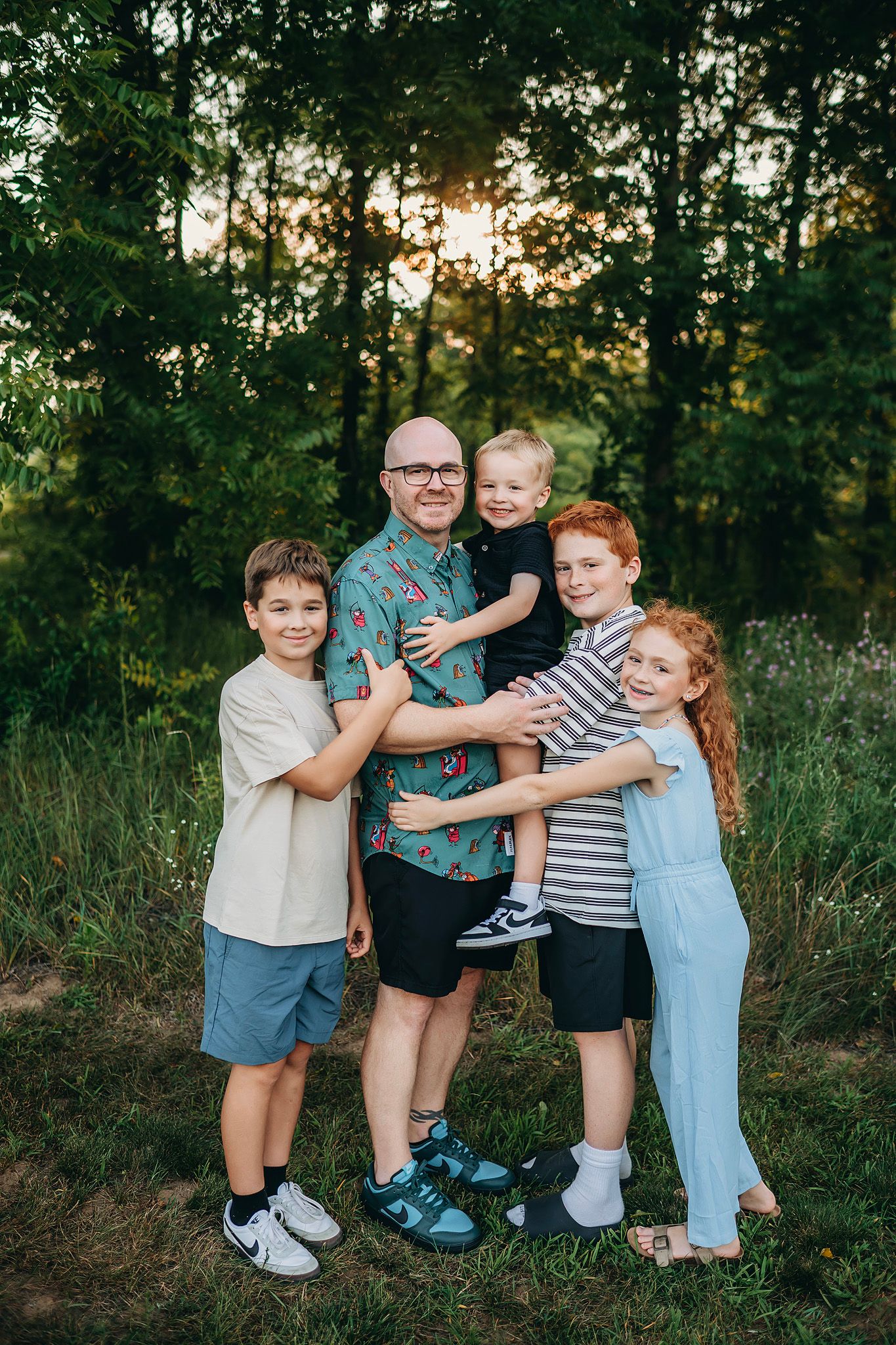 A father poses outdoors at sunset with four children hugging him, surrounded by lush green trees.