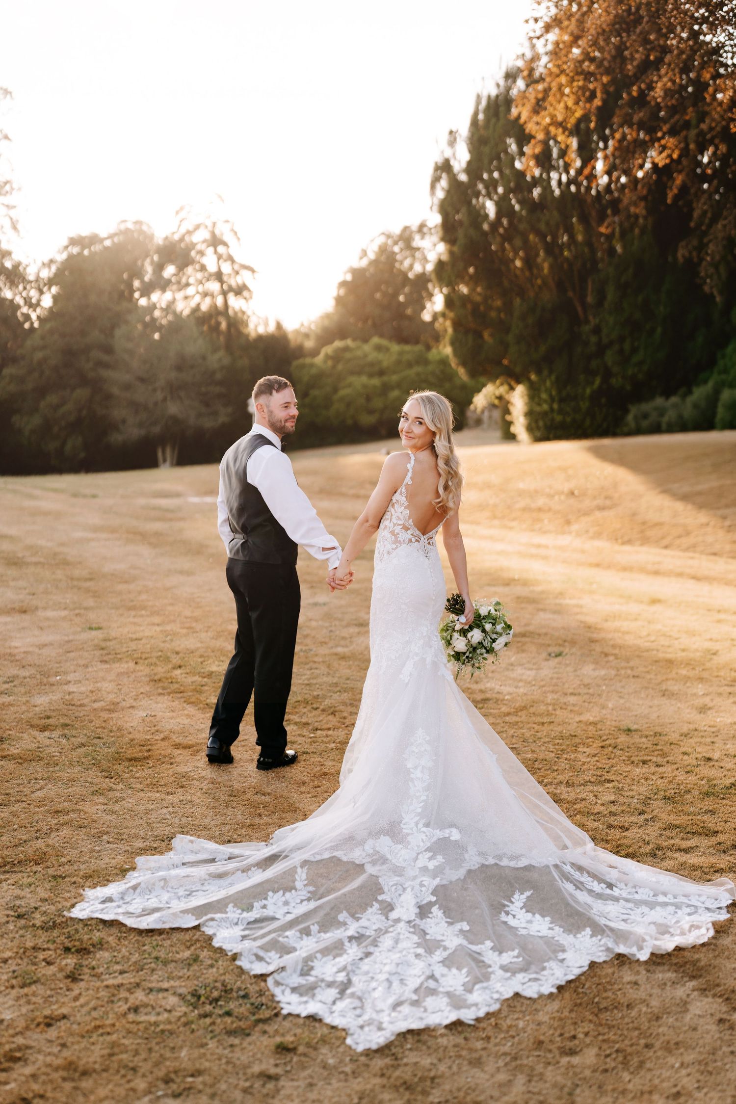 A couple in wedding attire walks along a stone path beside an elegant building with flowering shrubs and ivy-covered walls.