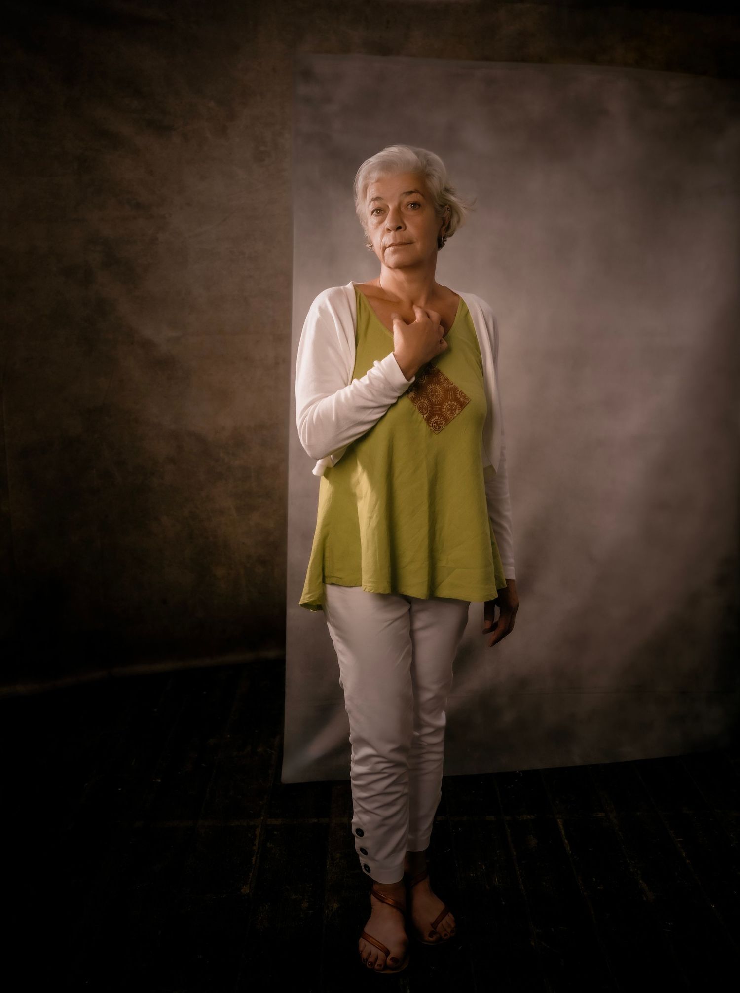 Elderly woman with short gray hair poses in green top, white cardigan and pants against dark studio backdrop.