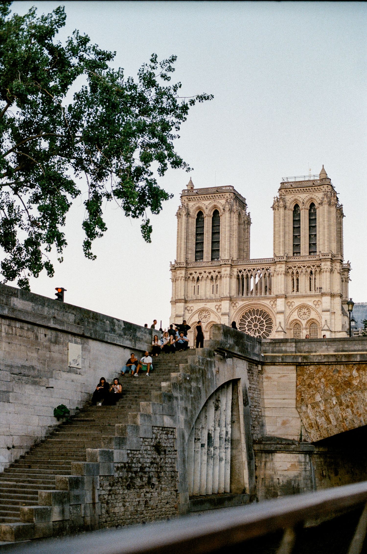 Notre-Dame Cathedral towers over a stone bridge along the Seine River in Paris, with visitors relaxing on the steps.