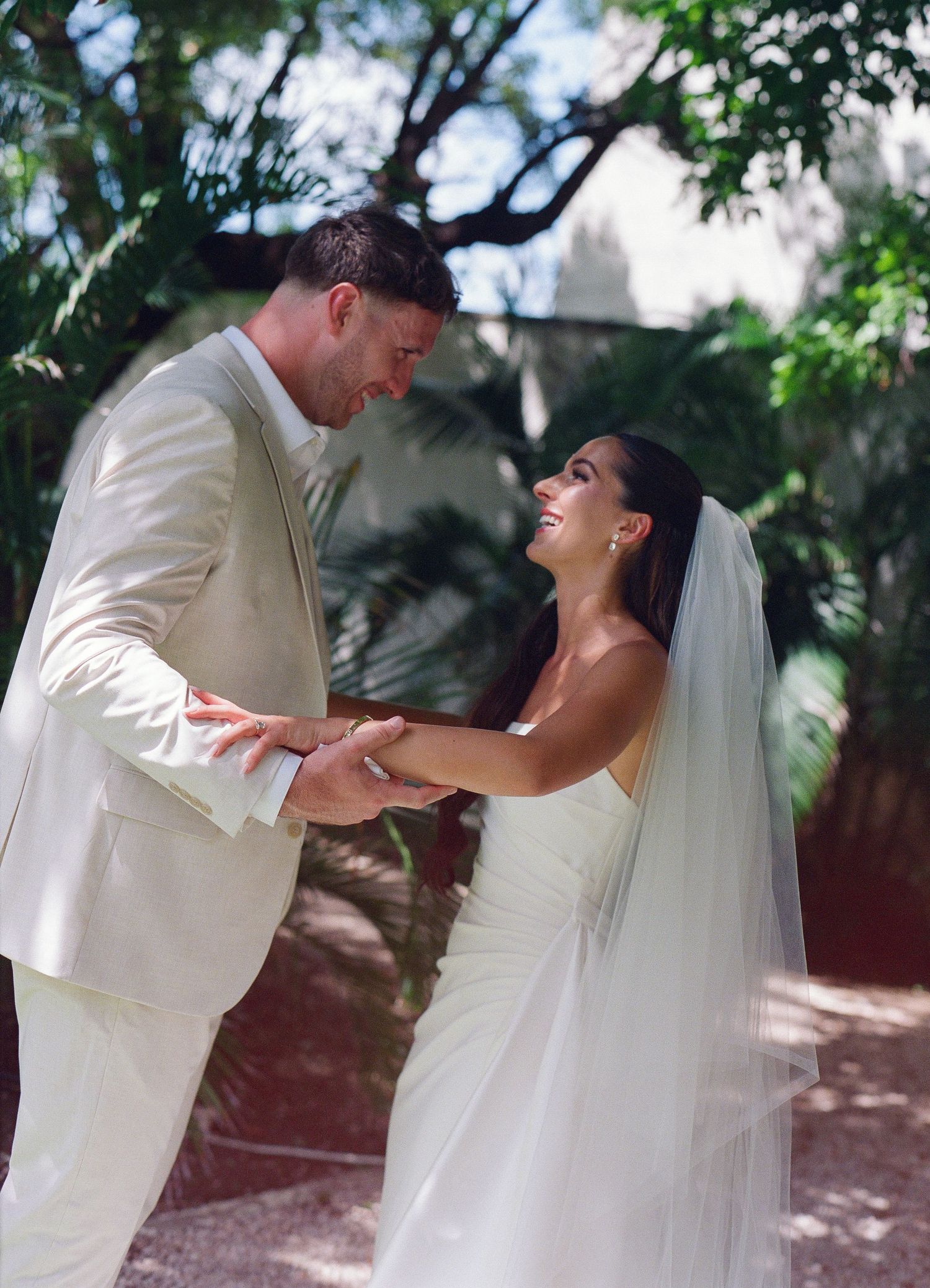 Bride and groom holding hands and smiling at each other outdoors in white wedding attire surrounded by lush greenery.