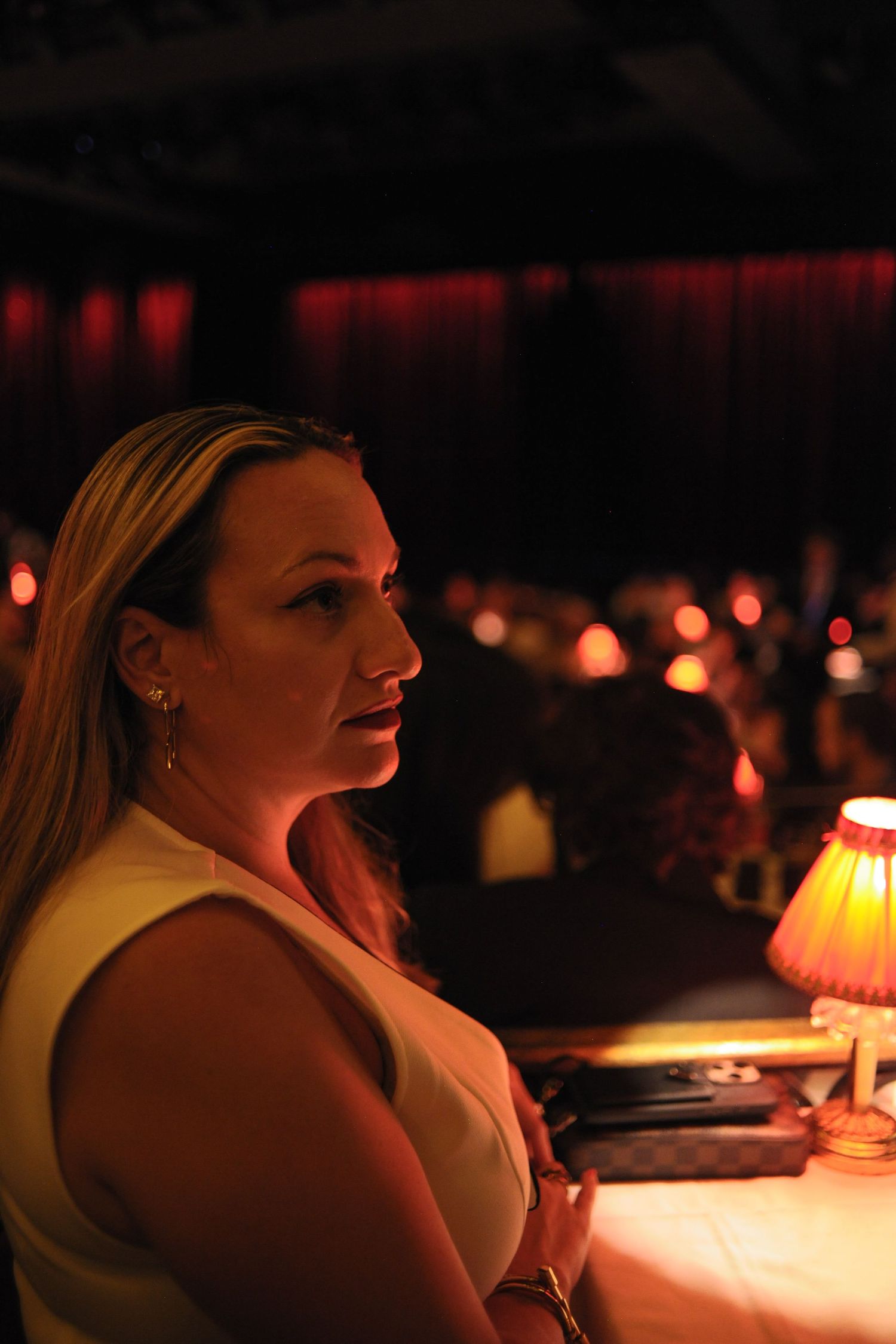 Blonde woman in white dress seated at candlelit table in dimly lit venue with red curtains.