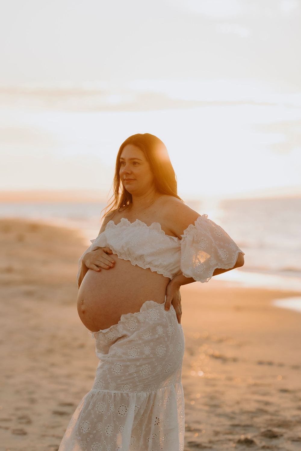 Pregnant woman in white lace dress cradling baby bump on beach at golden sunset.