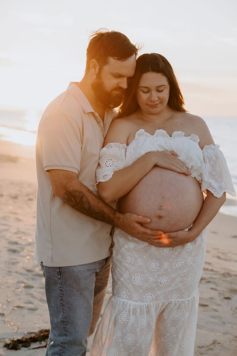 Couple tenderly cradling pregnant belly on sandy beach at golden sunset, woman in white lace dress.