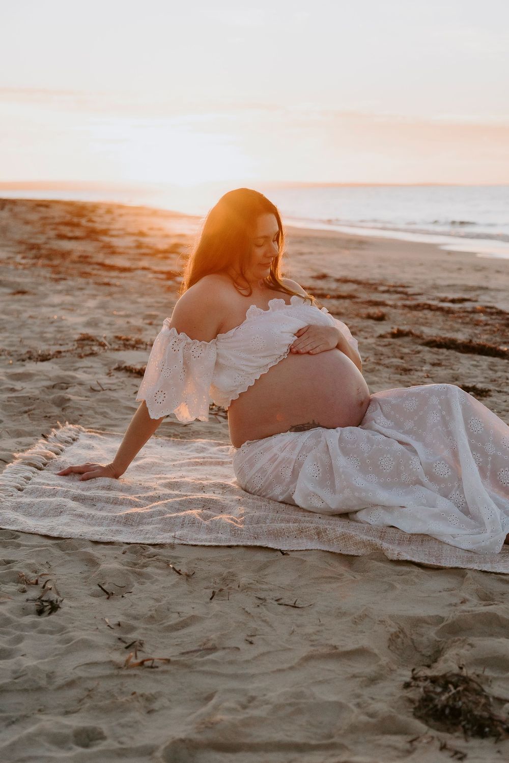 Pregnant woman in white lace dress sitting on beach blanket at golden sunset, cradling baby bump.